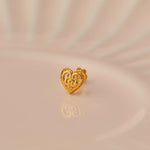 Close up of a heart shaped 18K gold nose pin placed on a soft peach surface highlighting its ornate detailing