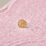 Close up of a textured round 18K gold nose pin placed on a soft pink background highlighting fine detailing