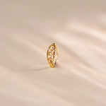 Close up of an 18K gold nose ring with intricate floral cutwork pattern, displayed upright on a soft beige fabric surface with warm natural lighting