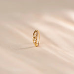 Close up of an 18K gold nose ring with intricate cutwork detailing standing upright on a soft beige fabric background with gentle light reflections