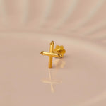 Close up of a cross shaped 18K gold nose pin placed on a soft peach background highlighting its polished finish
