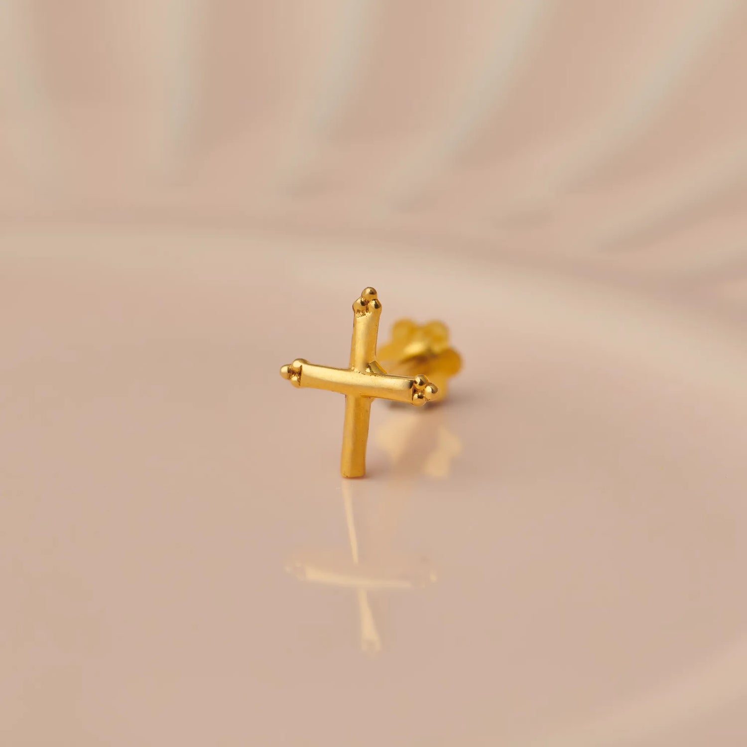 Close up of a cross shaped 18K gold nose pin placed on a soft peach background highlighting its polished finish