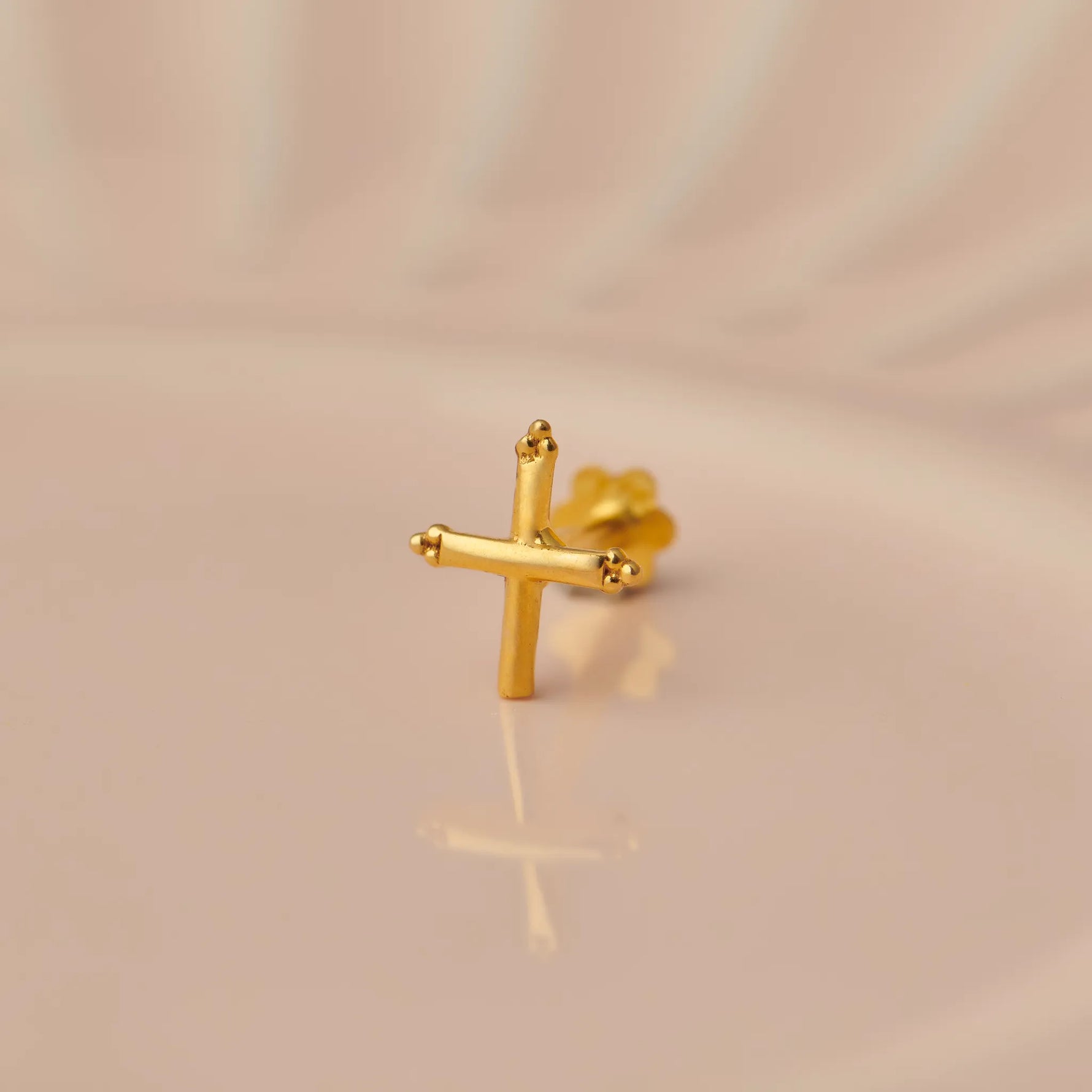 Close up of a cross shaped 18K gold nose pin placed on a soft peach background highlighting its polished finish