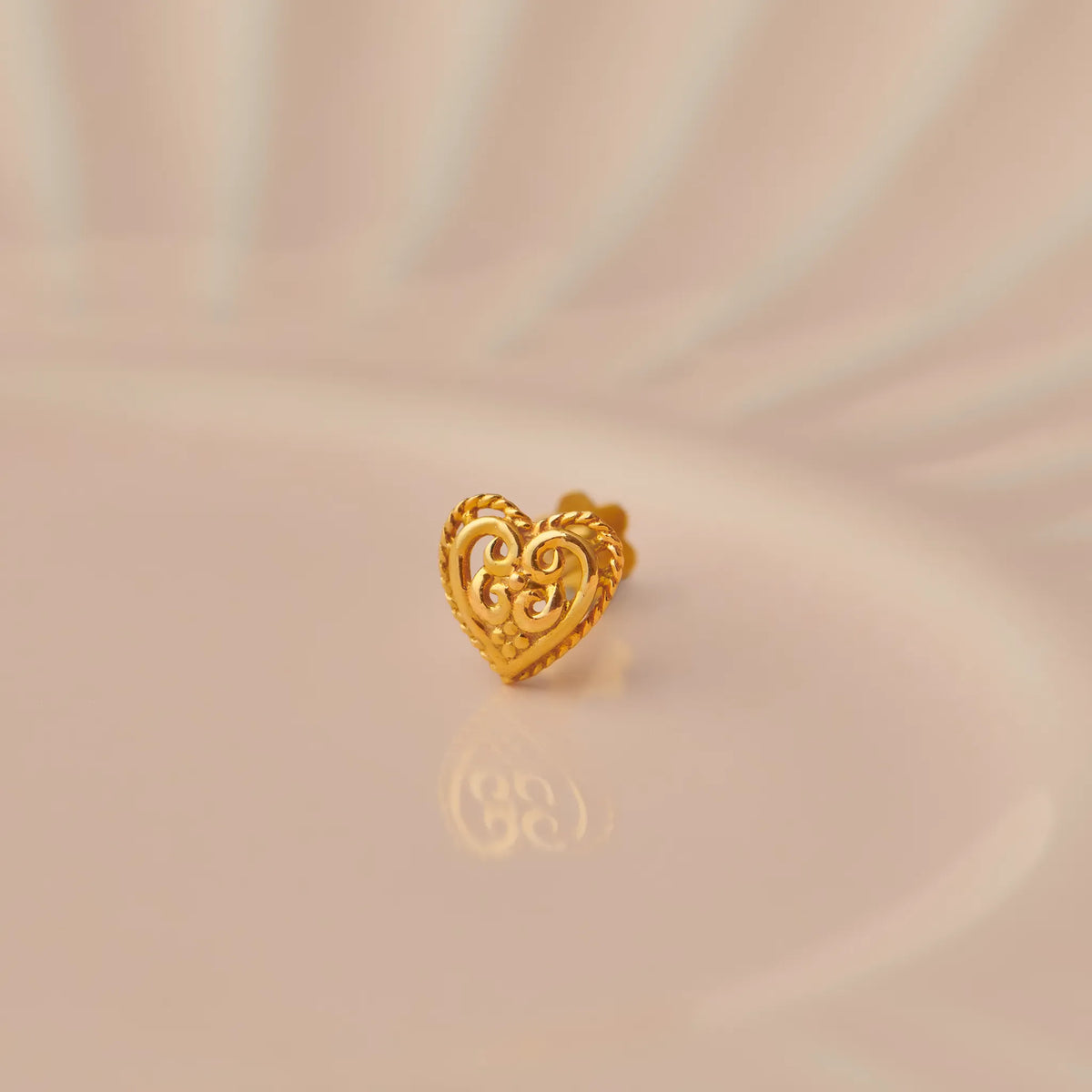 Close up of a heart shaped 18K gold nose pin placed on a soft peach surface highlighting its ornate detailing
