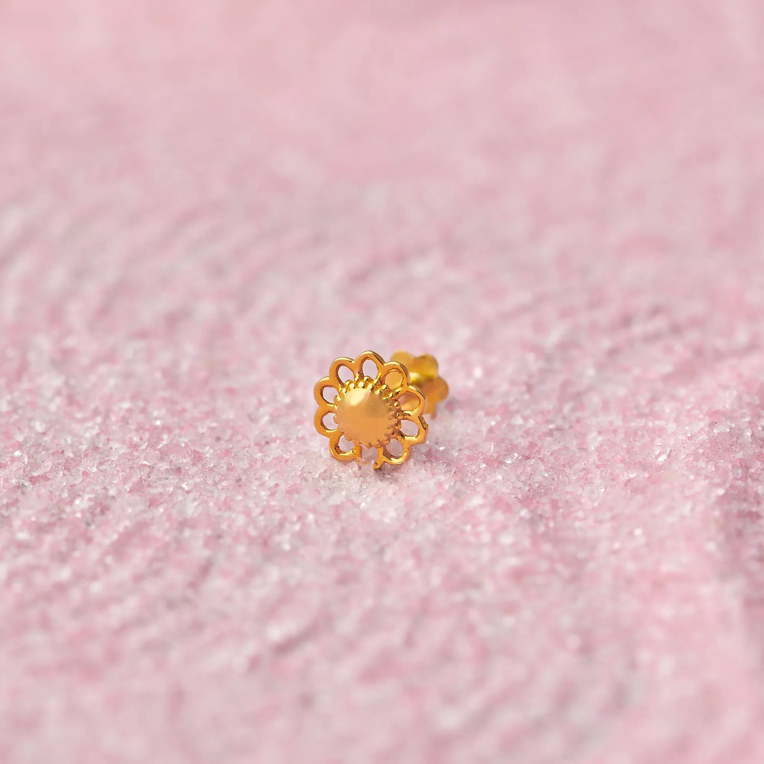 Close up of a floral inspired 18K gold nose pin placed on a soft pink textured background highlighting its intricate design