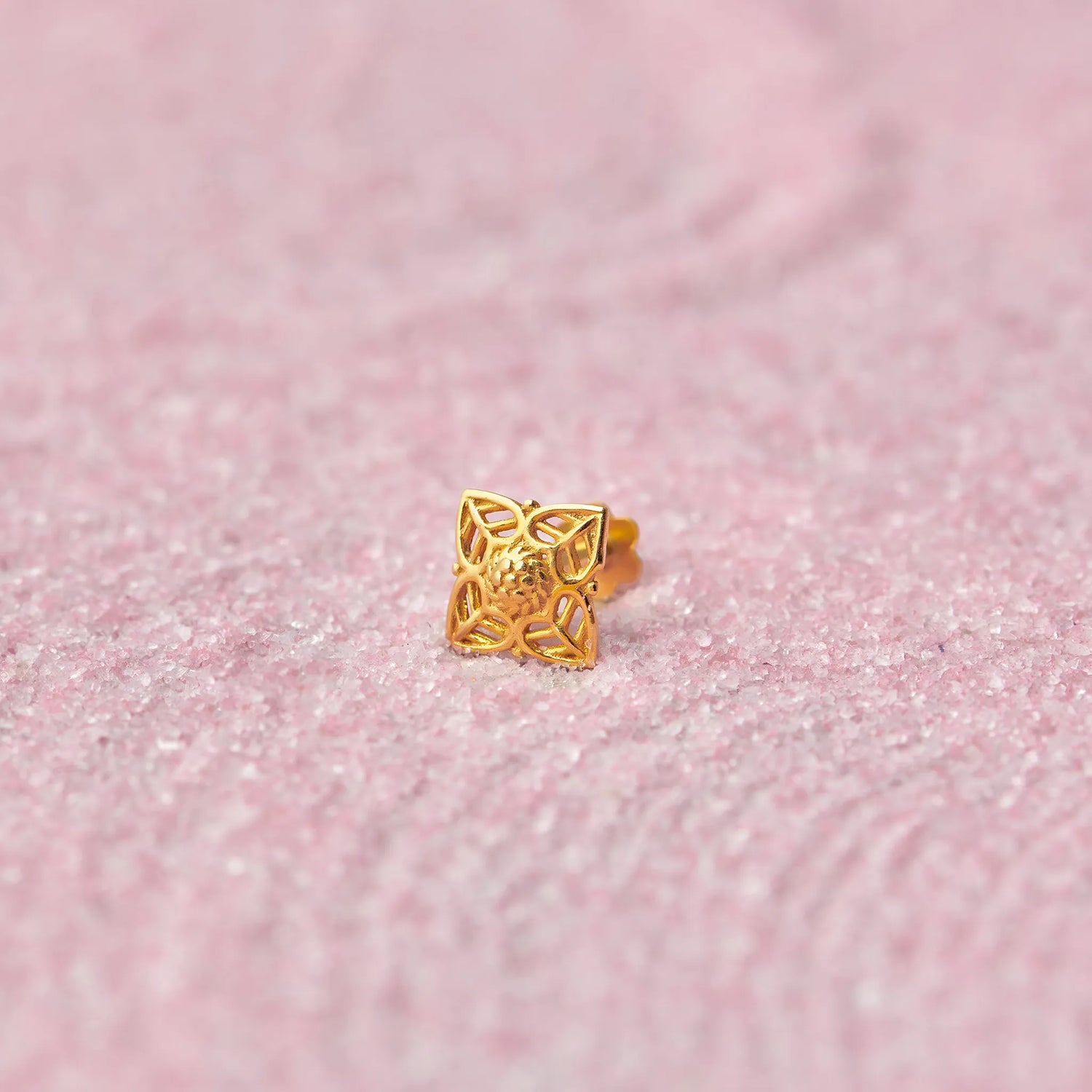 Close up of square floral inspired 18K gold nose pin placed on a soft pink textured background showcasing intricate cutwork detailing