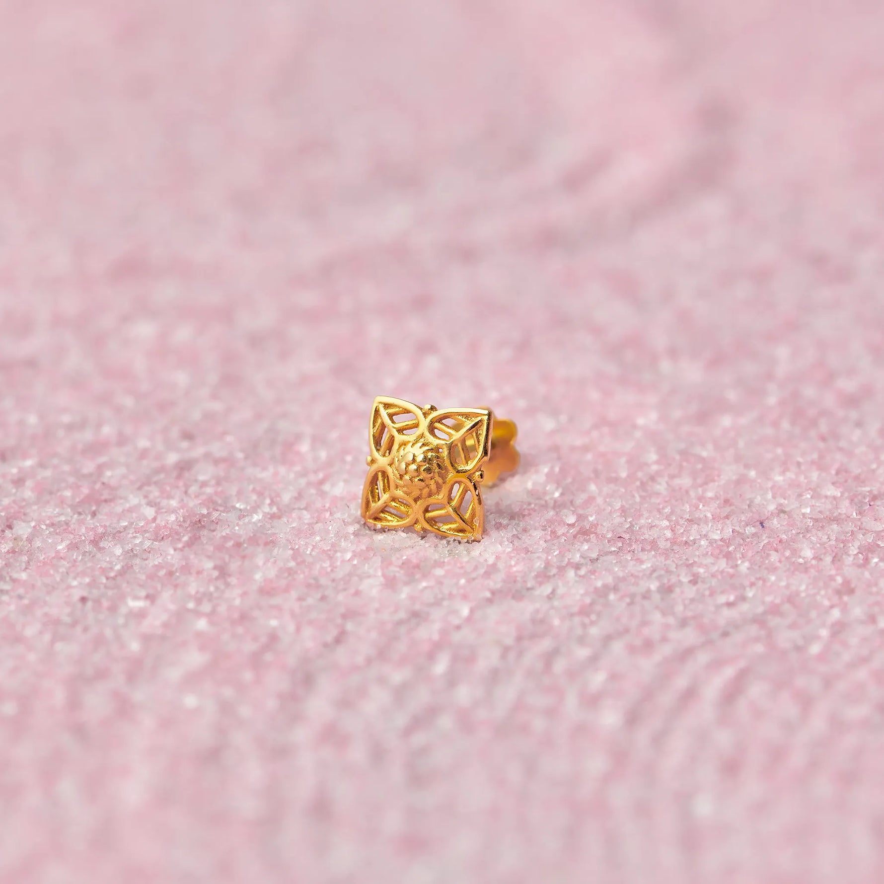 Close up of square floral inspired 18K gold nose pin placed on a soft pink textured background showcasing intricate cutwork detailing