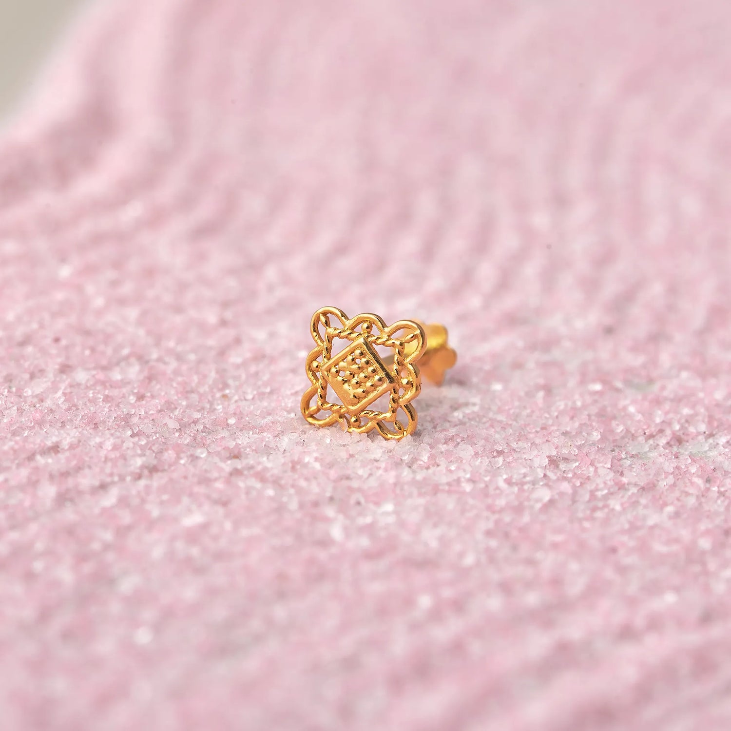 Close up of an intricate geometric design 18K gold nose pin placed on a soft pink textured background highlighting fine craftsmanship