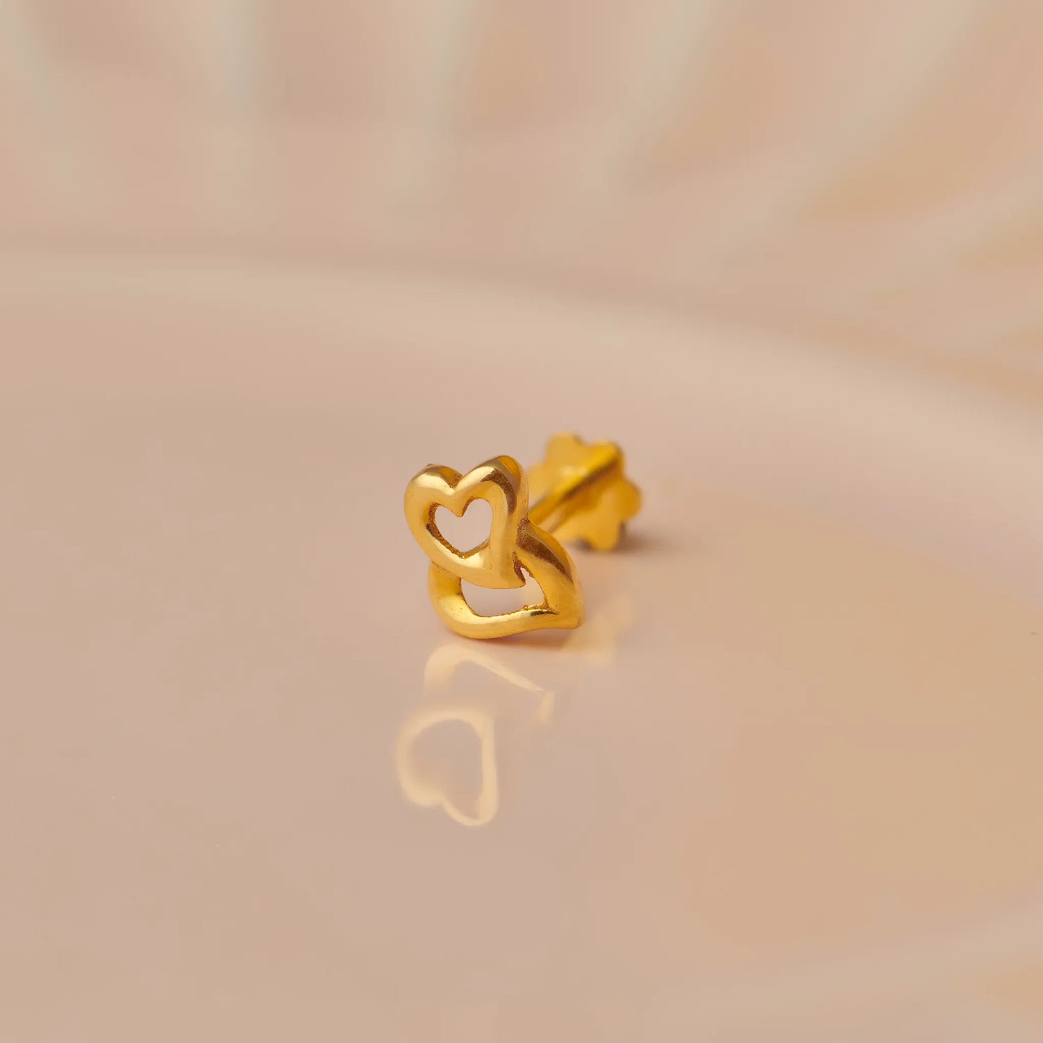 Close up of a delicate heart shaped 18K gold nose pin resting on a soft beige surface with subtle reflection