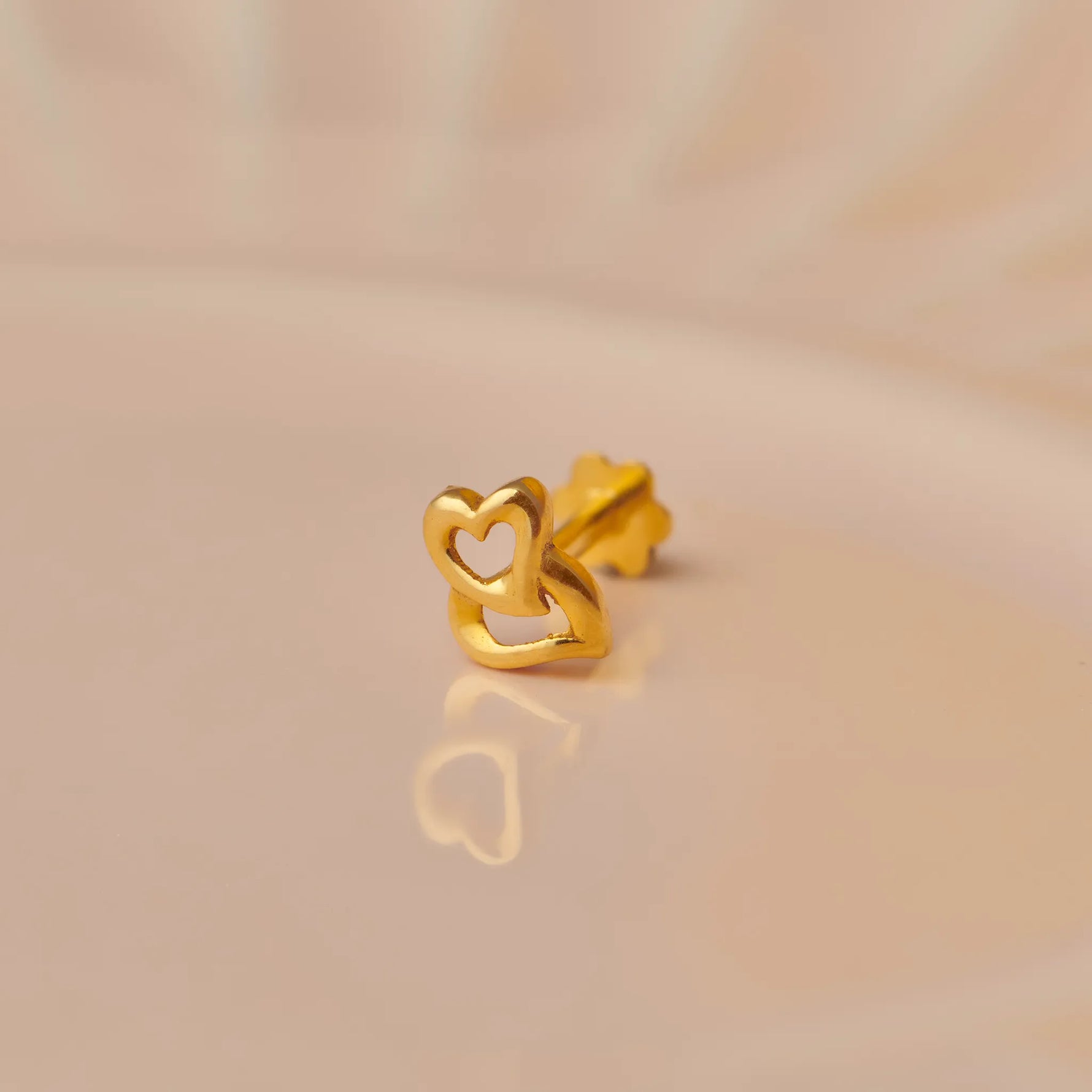 Close up of a delicate heart shaped 18K gold nose pin resting on a soft beige surface with subtle reflection