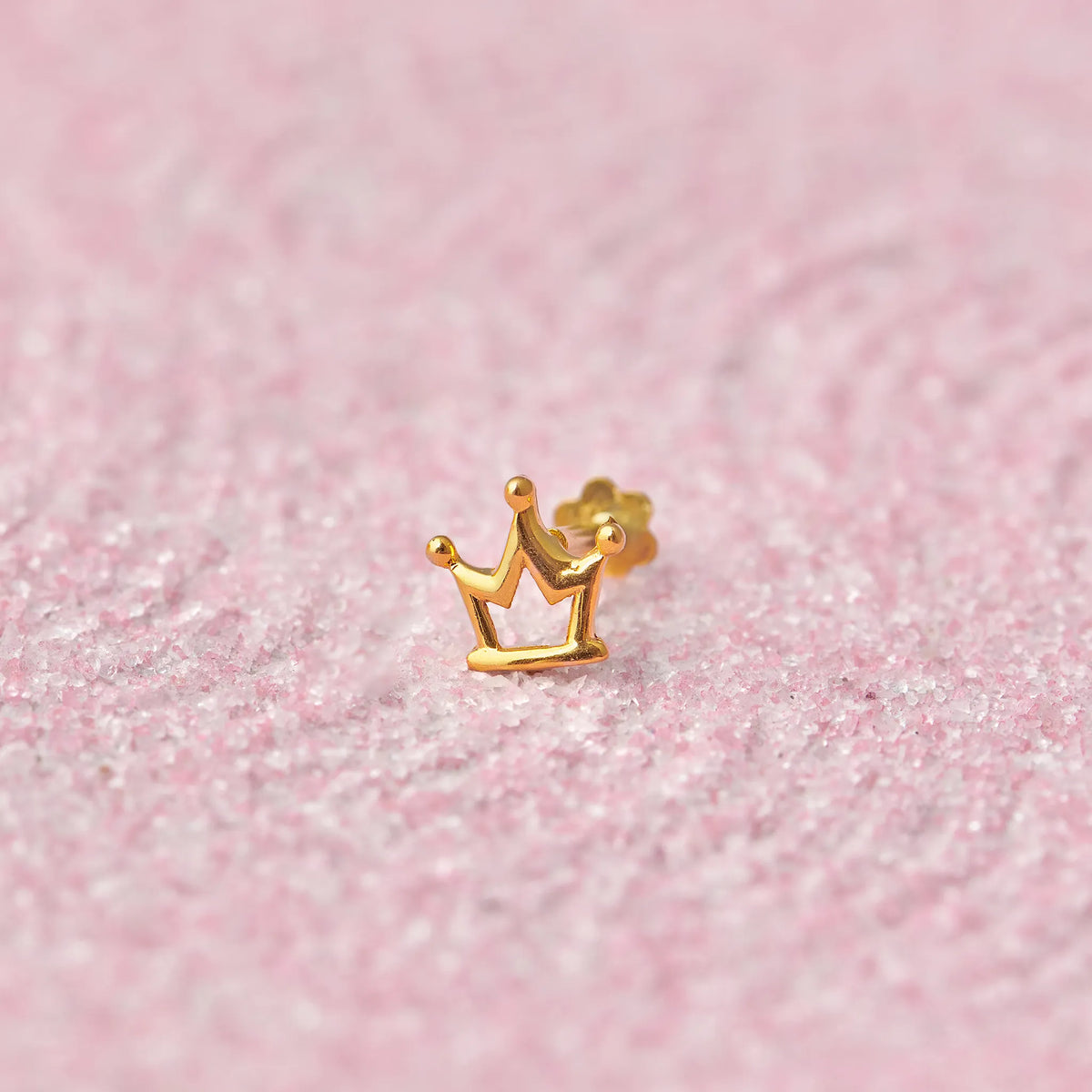 Close up of crown inspired gold nose pin placed on a soft pink textured background highlighting its royal design