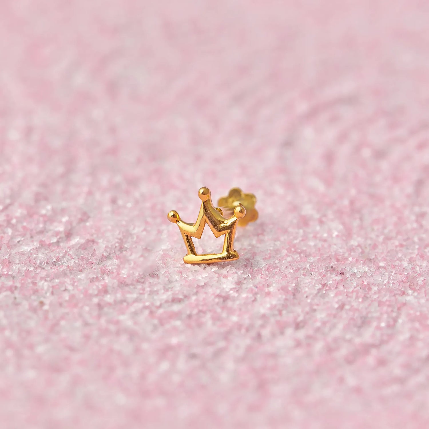 Close up of crown inspired gold nose pin placed on a soft pink textured background highlighting its royal design