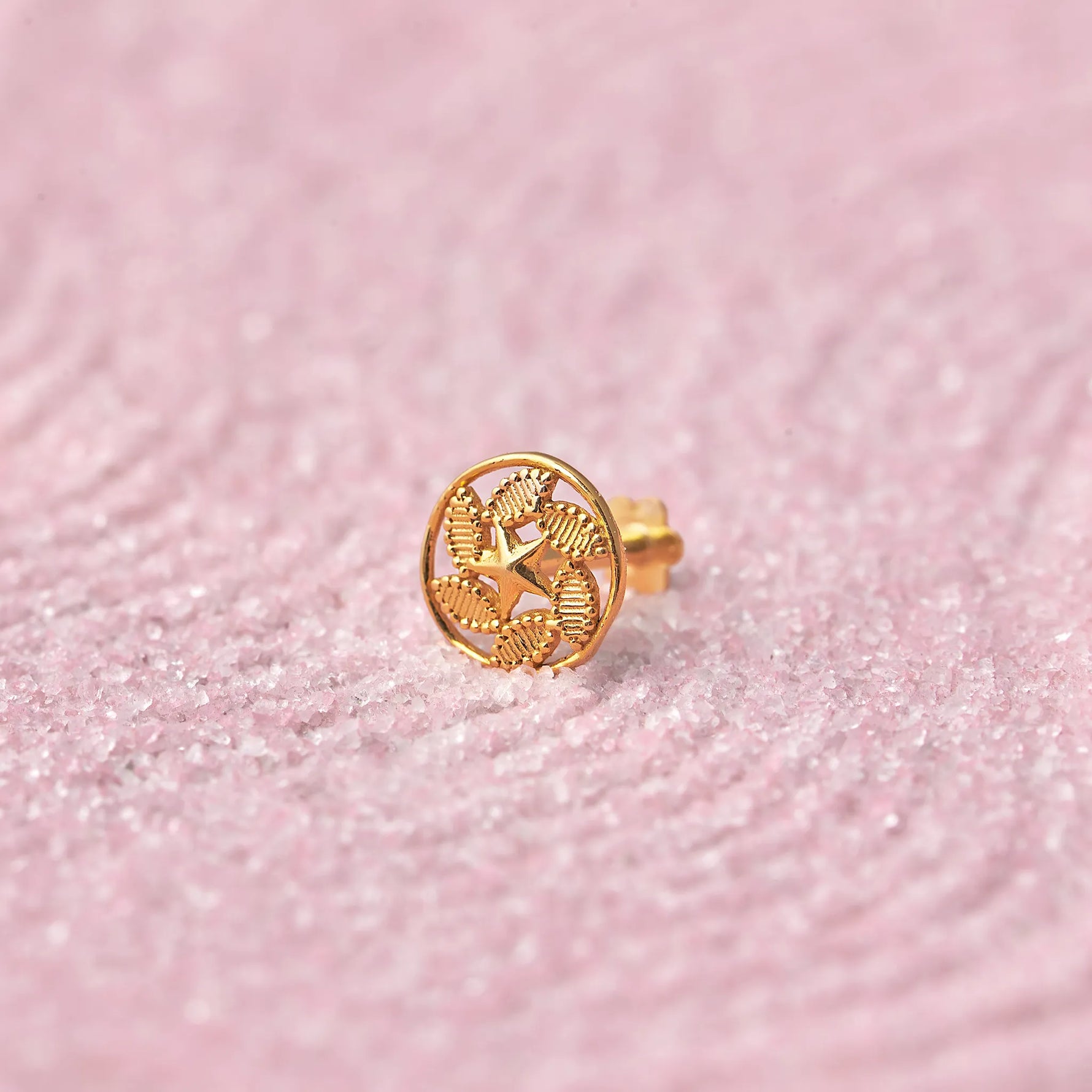 Close up of a round patterned 18K gold nose pin placed on a soft pink textured background highlighting detailed craftsmanship