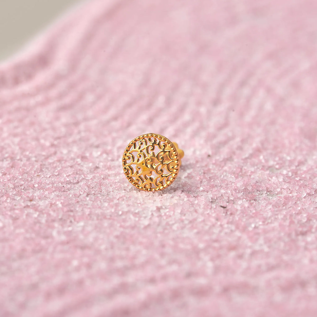 Close up of a textured round 18K gold nose pin placed on a soft pink background highlighting fine detailing