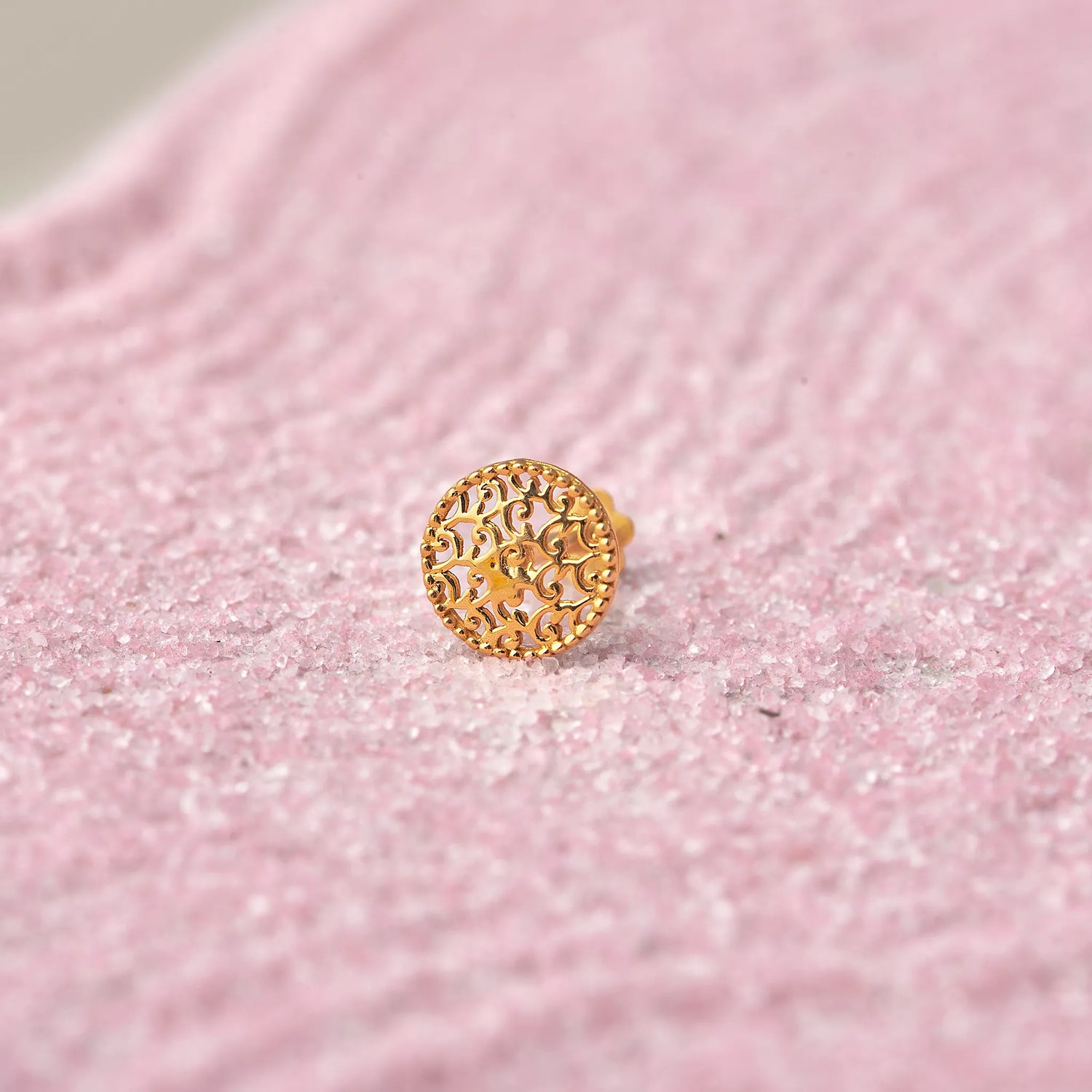 Close up of a textured round 18K gold nose pin placed on a soft pink background highlighting fine detailing