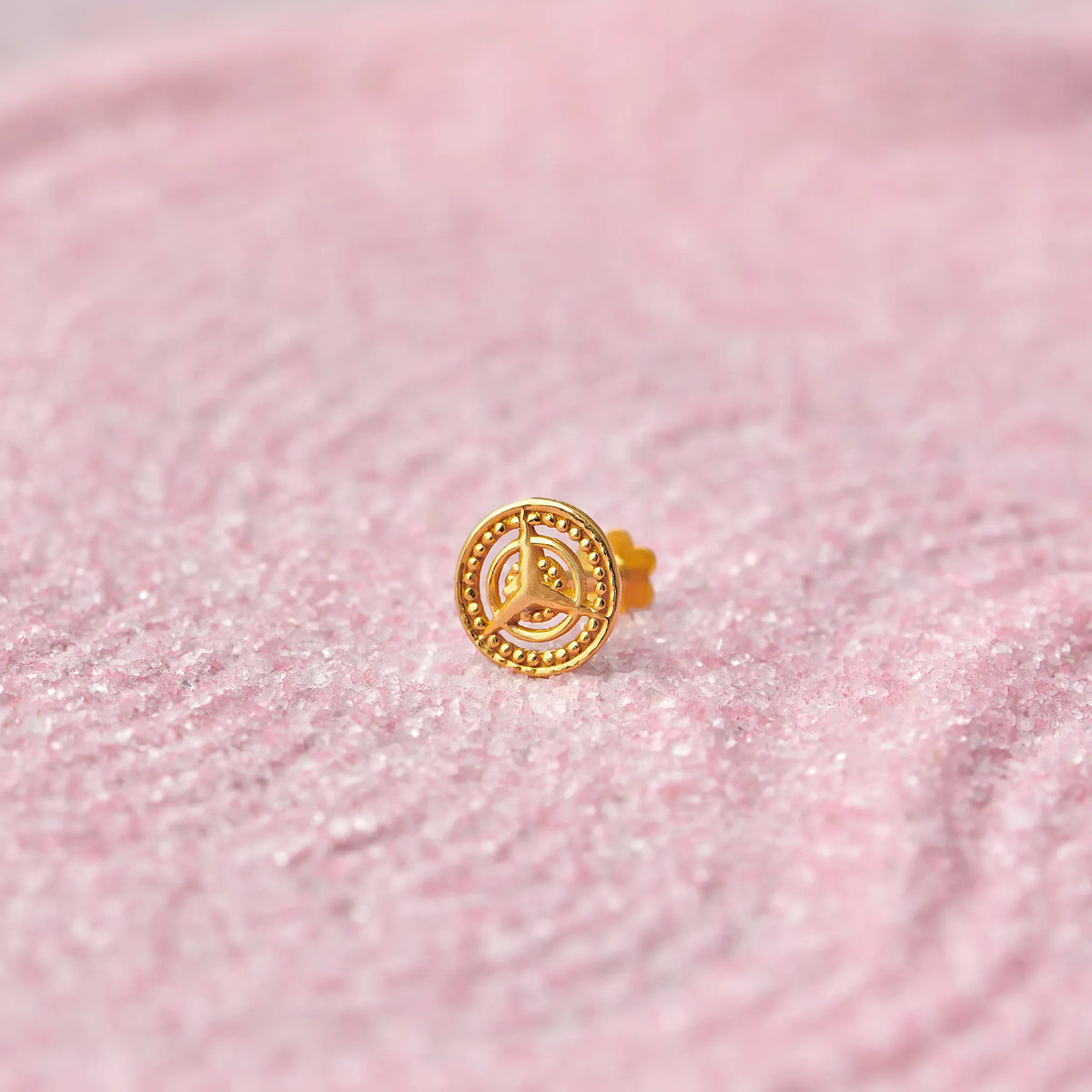 Close up view of a round 18K gold nose pin with spiral motif placed on a soft pink textured surface highlighting fine craftsmanship