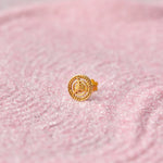 Close up view of a round 18K gold nose pin with spiral motif placed on a soft pink textured surface highlighting fine craftsmanship