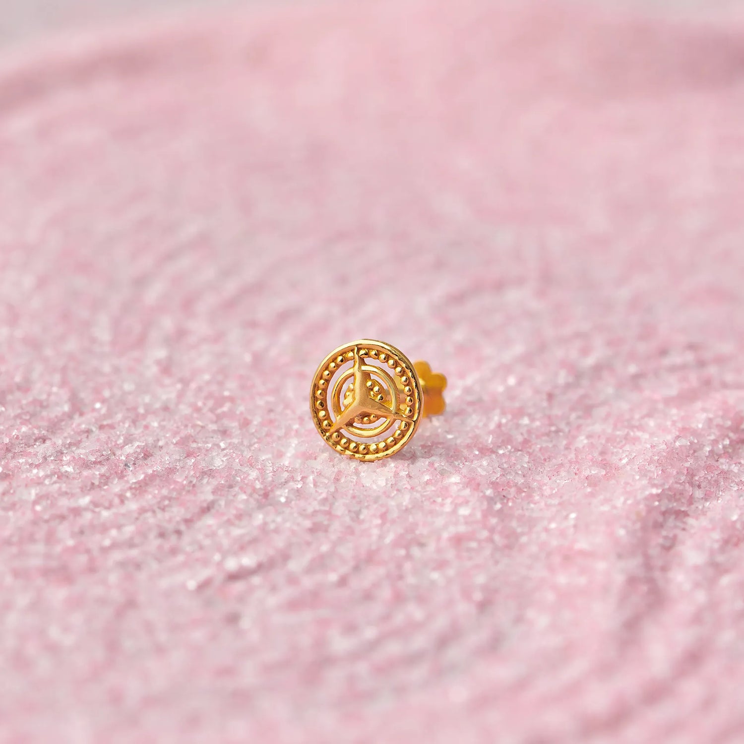 Close up view of a round 18K gold nose pin with spiral motif placed on a soft pink textured surface highlighting fine craftsmanship