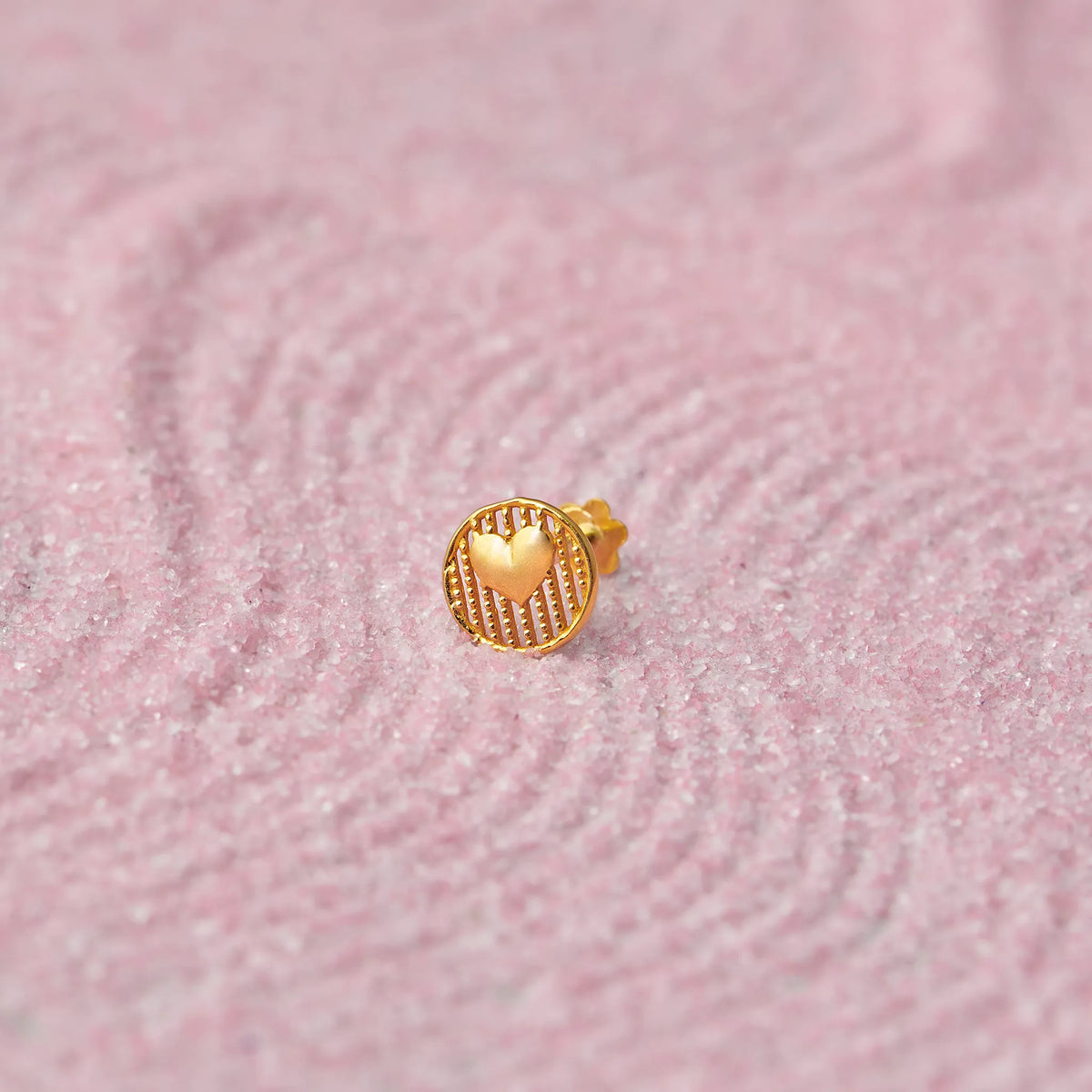 Close up of textured round heart inspired 18K gold nose pin placed on a soft pink fabric background highlighting fine surface detailing