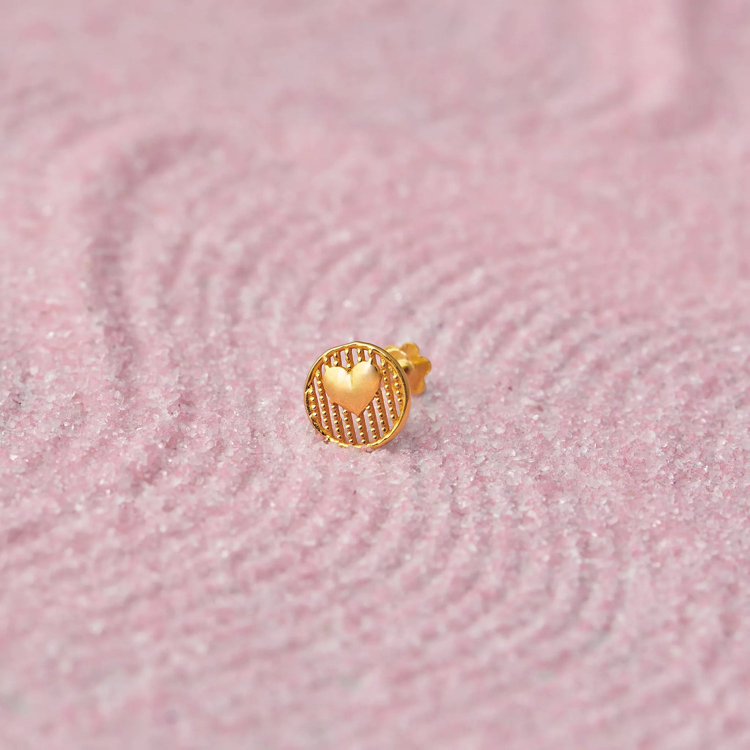 Close up of textured round heart inspired 18K gold nose pin placed on a soft pink fabric background highlighting fine surface detailing