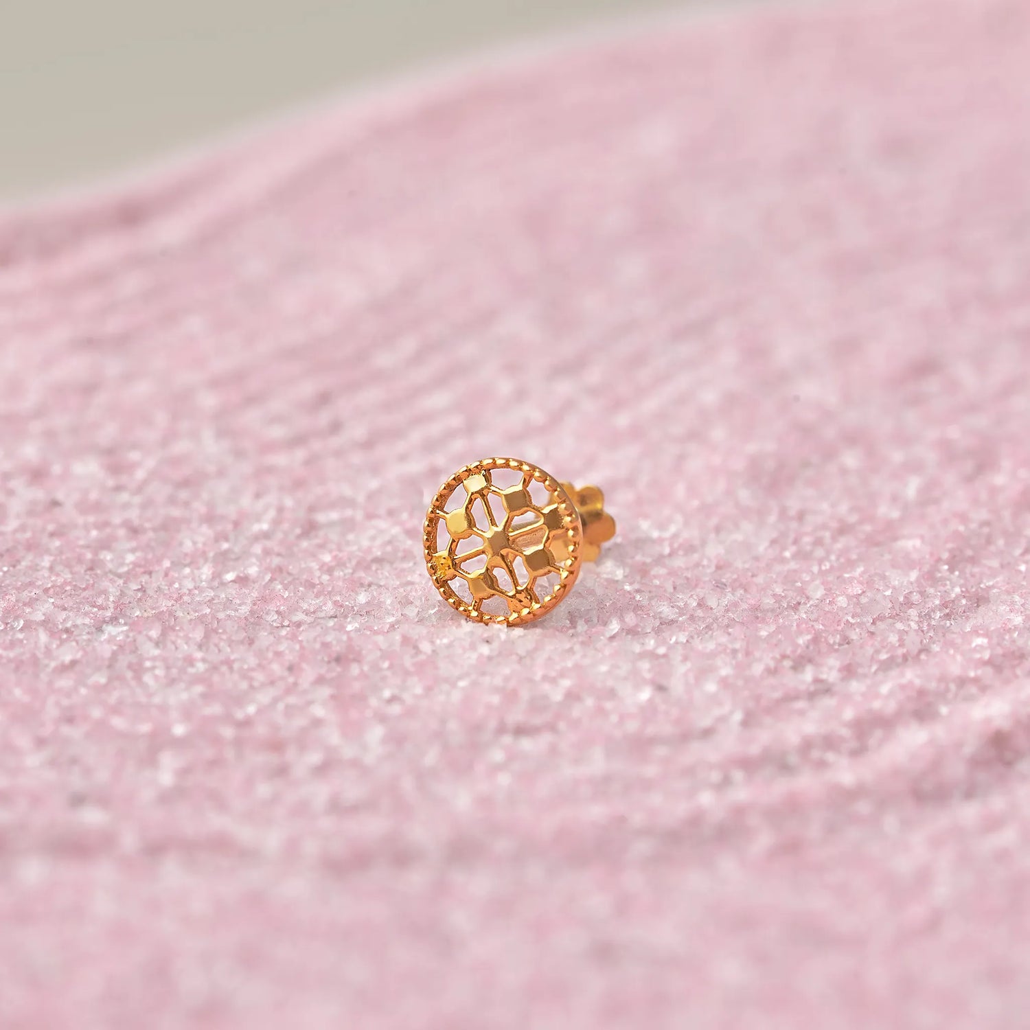 Close up view of a circular floral 18K gold nose pin placed on a soft pink textured surface highlighting intricate cutwork design