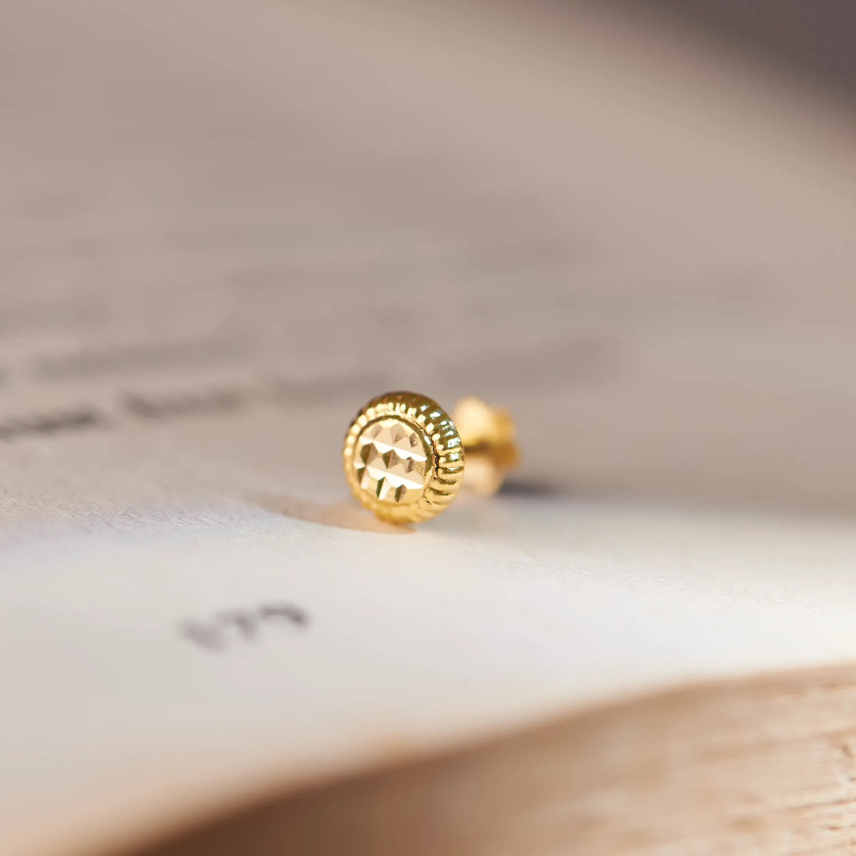 Close up of round textured 18K gold nose pin placed on an open book surface highlighting the polished finish and intricate edge detailing