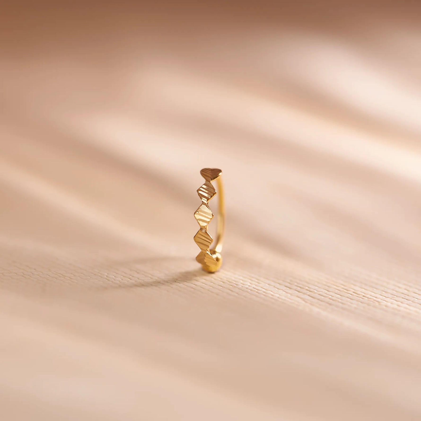 Close up of zigzag patterned 18K gold nose ring standing upright on a soft beige fabric background with natural light shadows