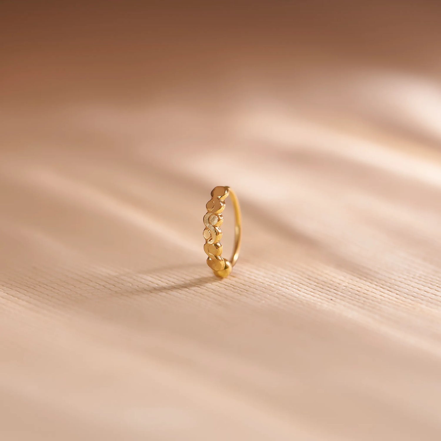 Close up of zigzag patterned 18K gold nose ring standing upright on a soft beige fabric background with gentle light reflections