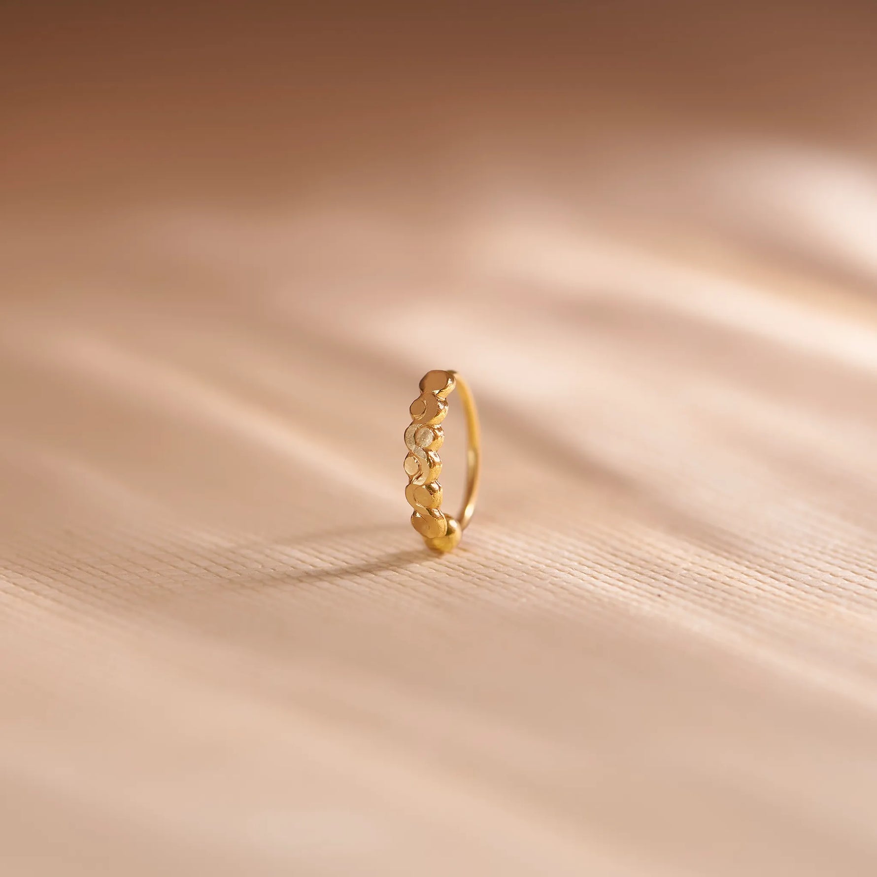 Close up of zigzag patterned 18K gold nose ring standing upright on a soft beige fabric background with gentle light reflections