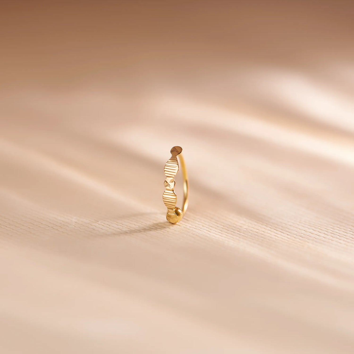 Close up of zigzag patterned 18K gold nose ring standing upright on a soft beige fabric background with warm natural light