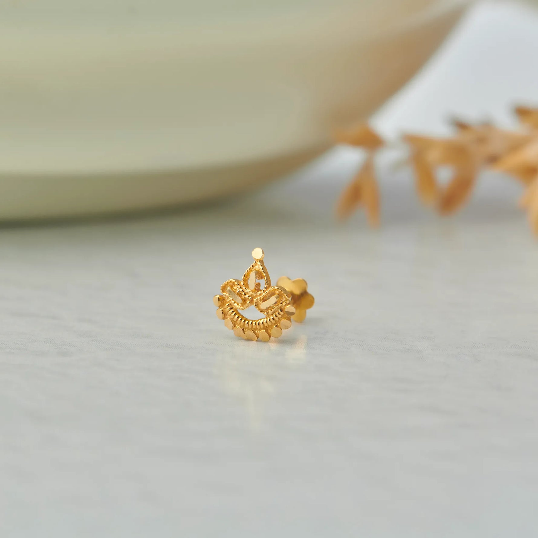 Close up of crown inspired 18K gold nose pin placed on a soft white surface with subtle decorative elements in the background highlighting regal detailing and fine craftsmanship