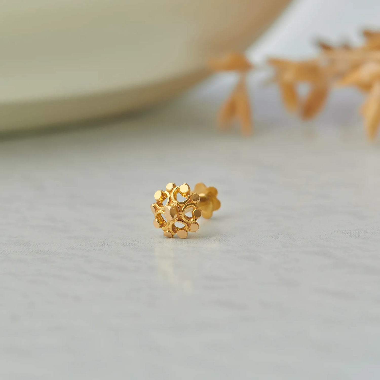 Close up of floral inspired 18K gold nose pin placed on a light textured surface with soft decor elements in the background