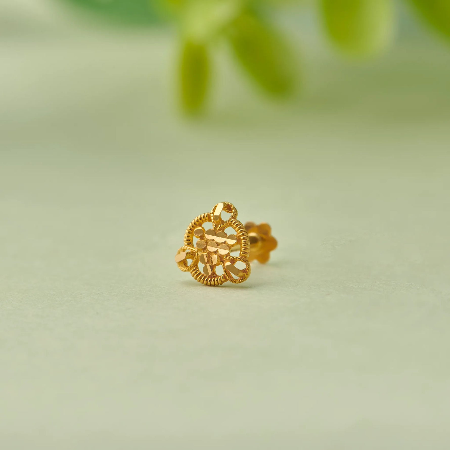 Close up of heart shaped 18K gold nose pin with intricate cutwork design placed on a soft green background with natural leaf accents