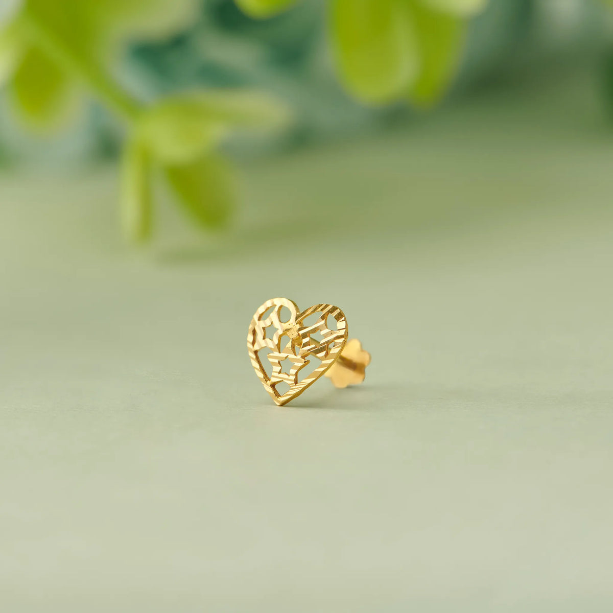 Close up of heart shaped filigree 18K gold nose pin placed on a soft pastel green surface with a blurred leafy background highlighting delicate openwork detailing