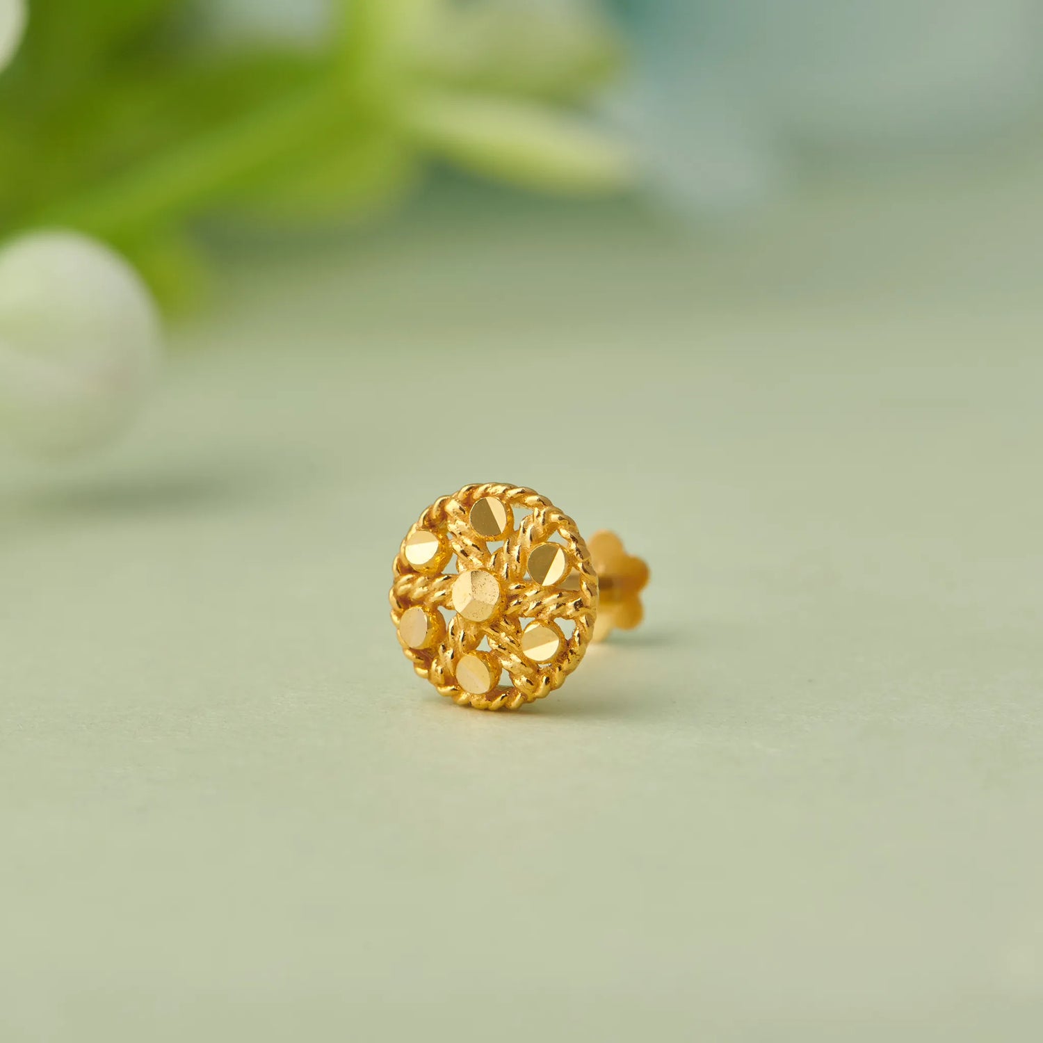 Close up of round floral inspired 18K gold nose pin placed on a soft green background with blurred leaf accents