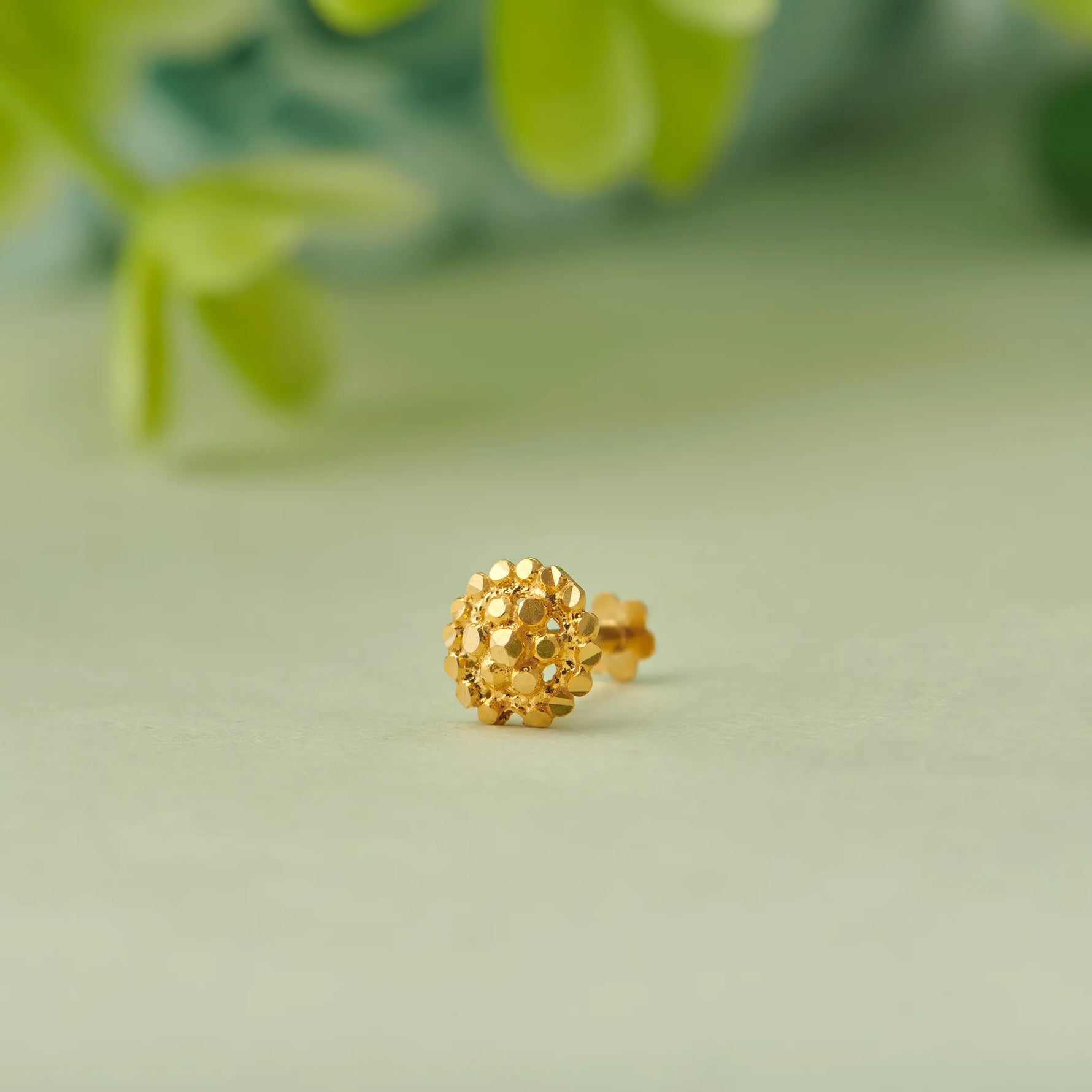 Close up of textured round floral inspired 18K gold nose pin placed on a soft green surface with blurred leaf accents