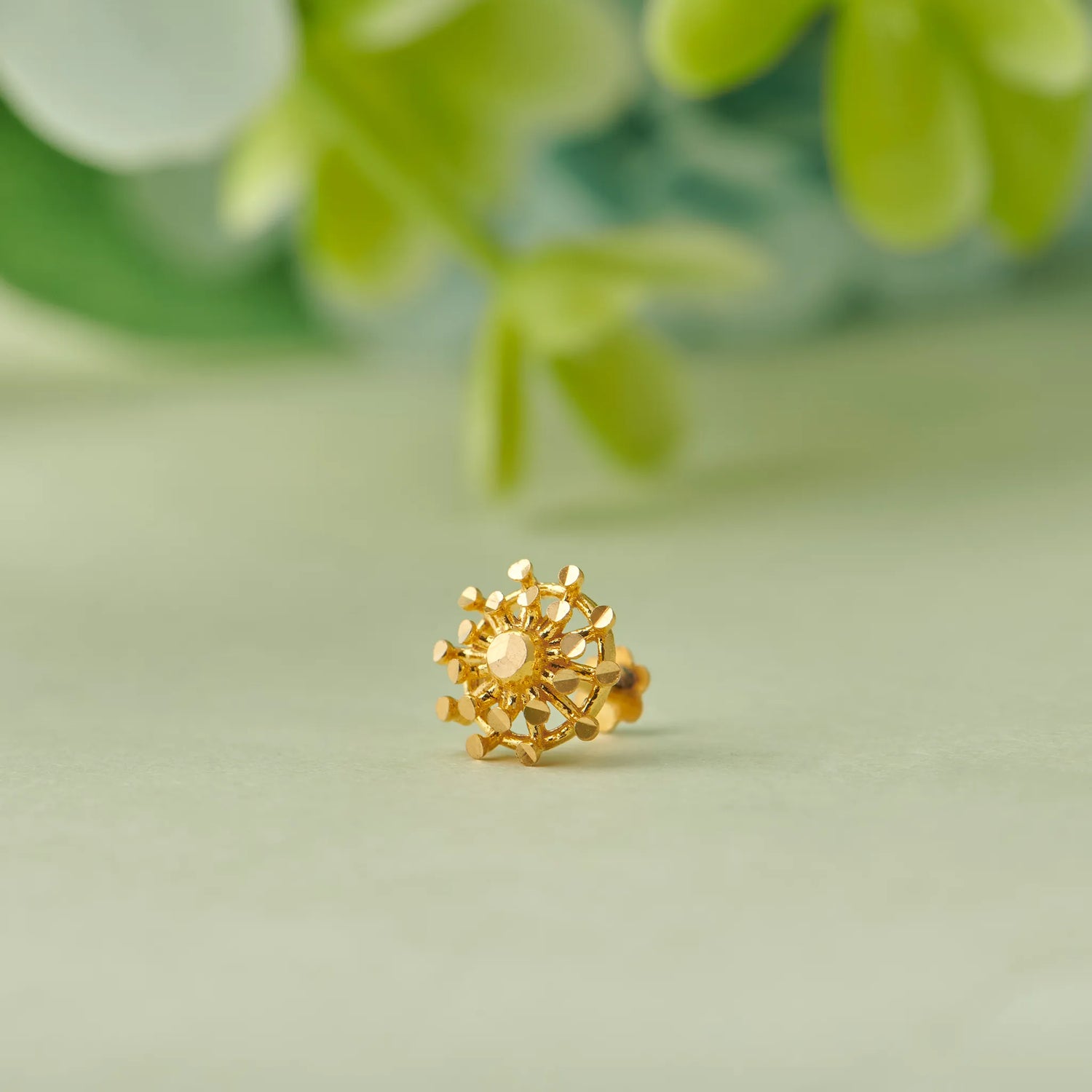 Close up of floral inspired 18K gold nose pin placed on a soft pastel green surface with blurred leafy background highlighting intricate craftsmanship