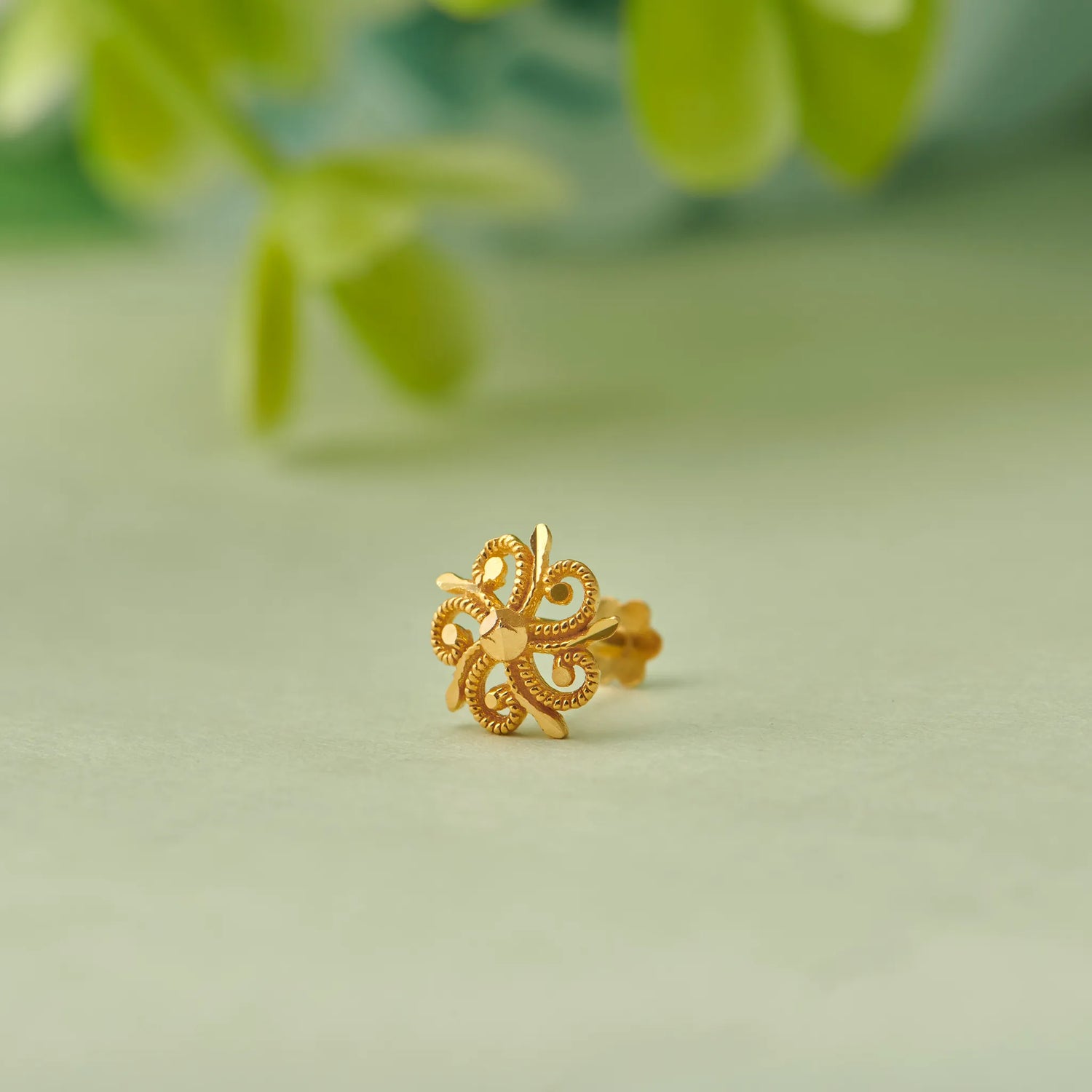 Close up of swirl inspired 18K gold nose pin placed on a soft green surface with blurred natural leaves in the background