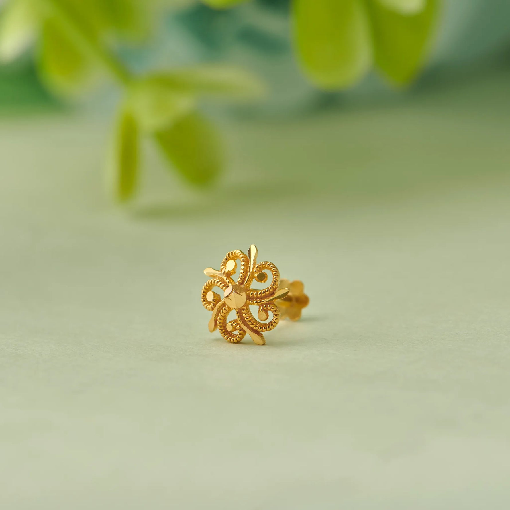 Close up of swirl inspired 18K gold nose pin placed on a soft green surface with blurred natural leaves in the background