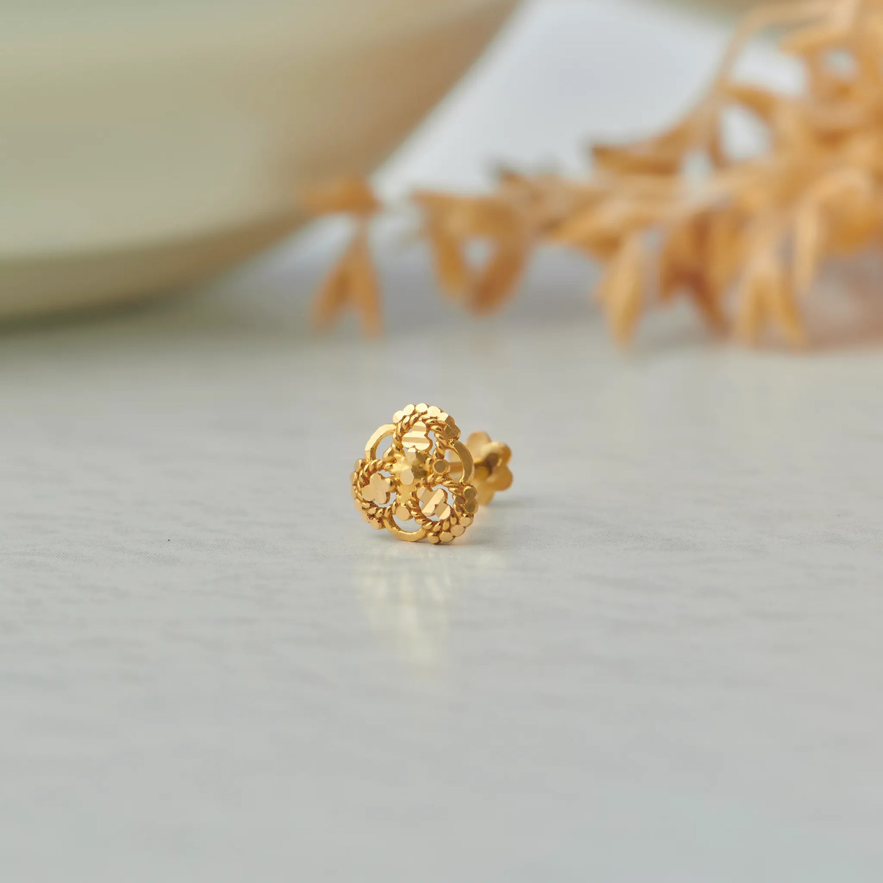 Close up of floral inspired 18K gold nose pin placed on a soft neutral surface with dried botanical accents highlighting fine craftsmanship