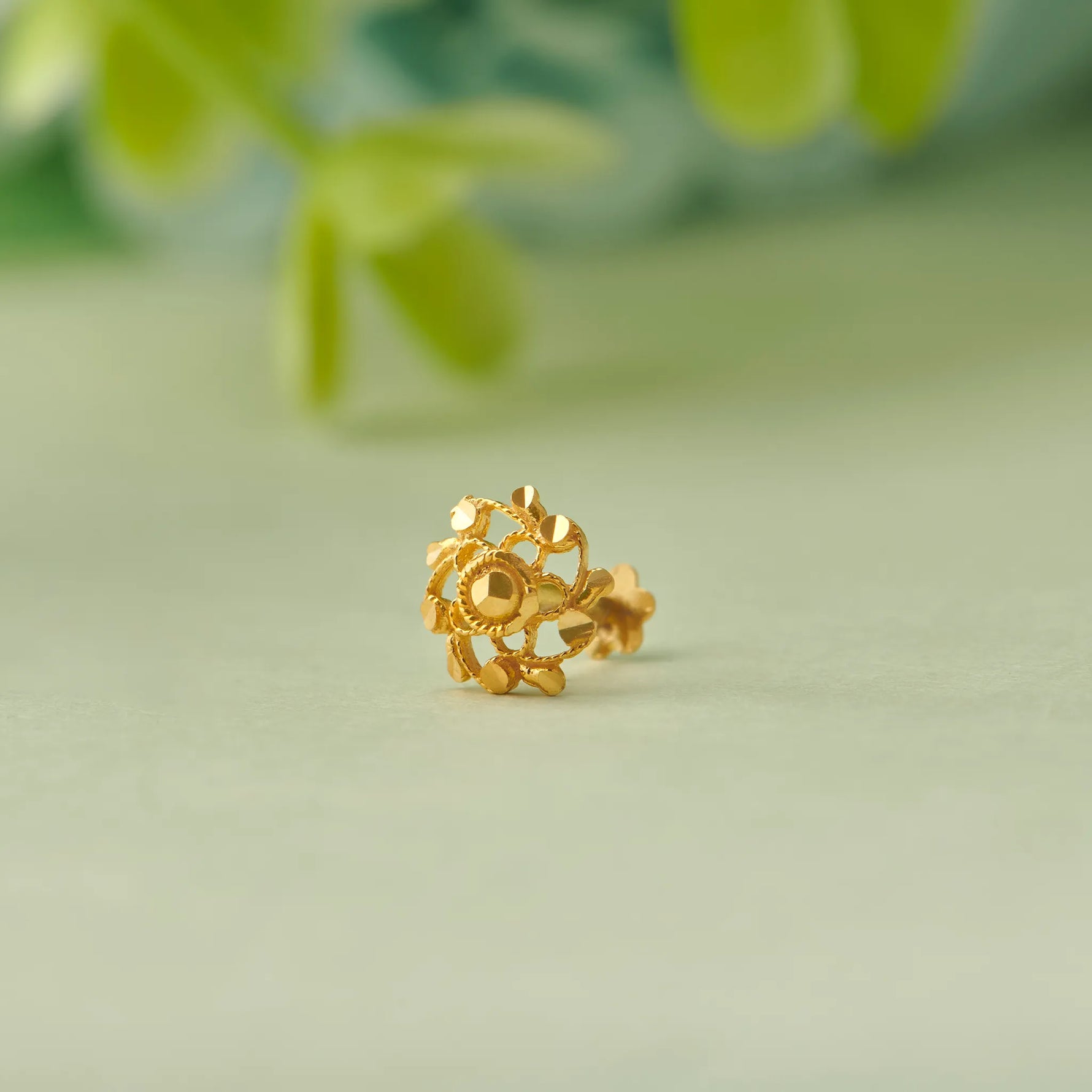 Close up of floral gold nose pin with green and red stones placed on a soft green background with blurred leaves