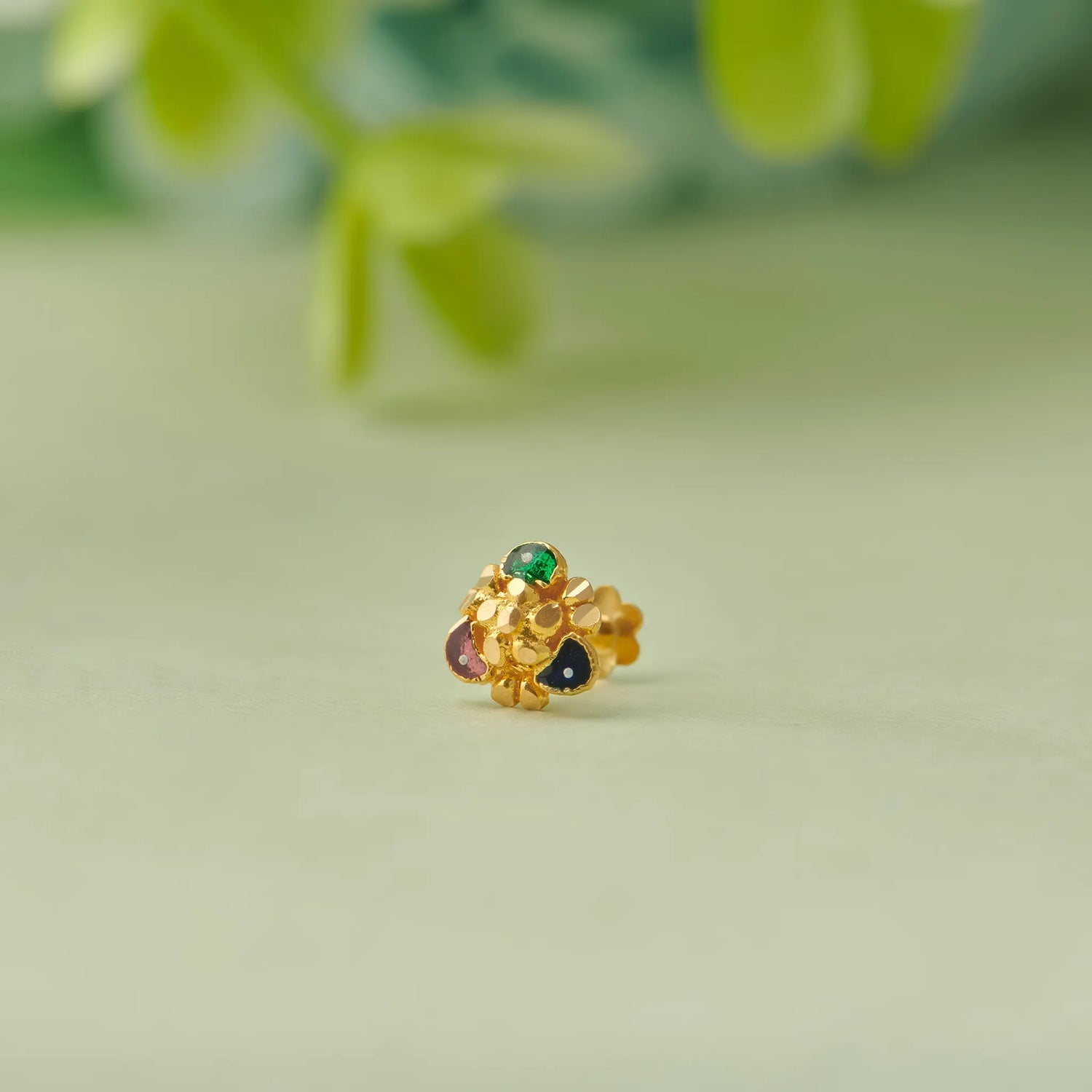 Close up of floral gold nose pin with green and red stones placed on a soft green background with blurred leaves