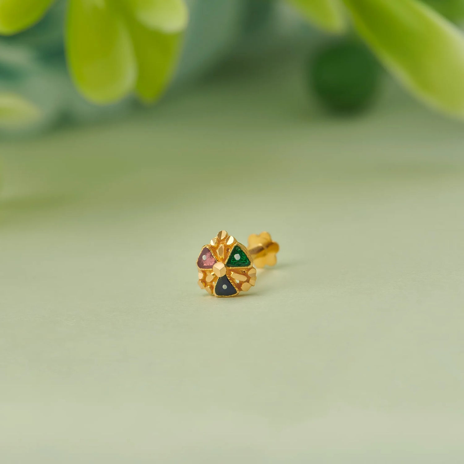 Close up of floral gold nose pin with green and red stone detailing placed on a soft green background