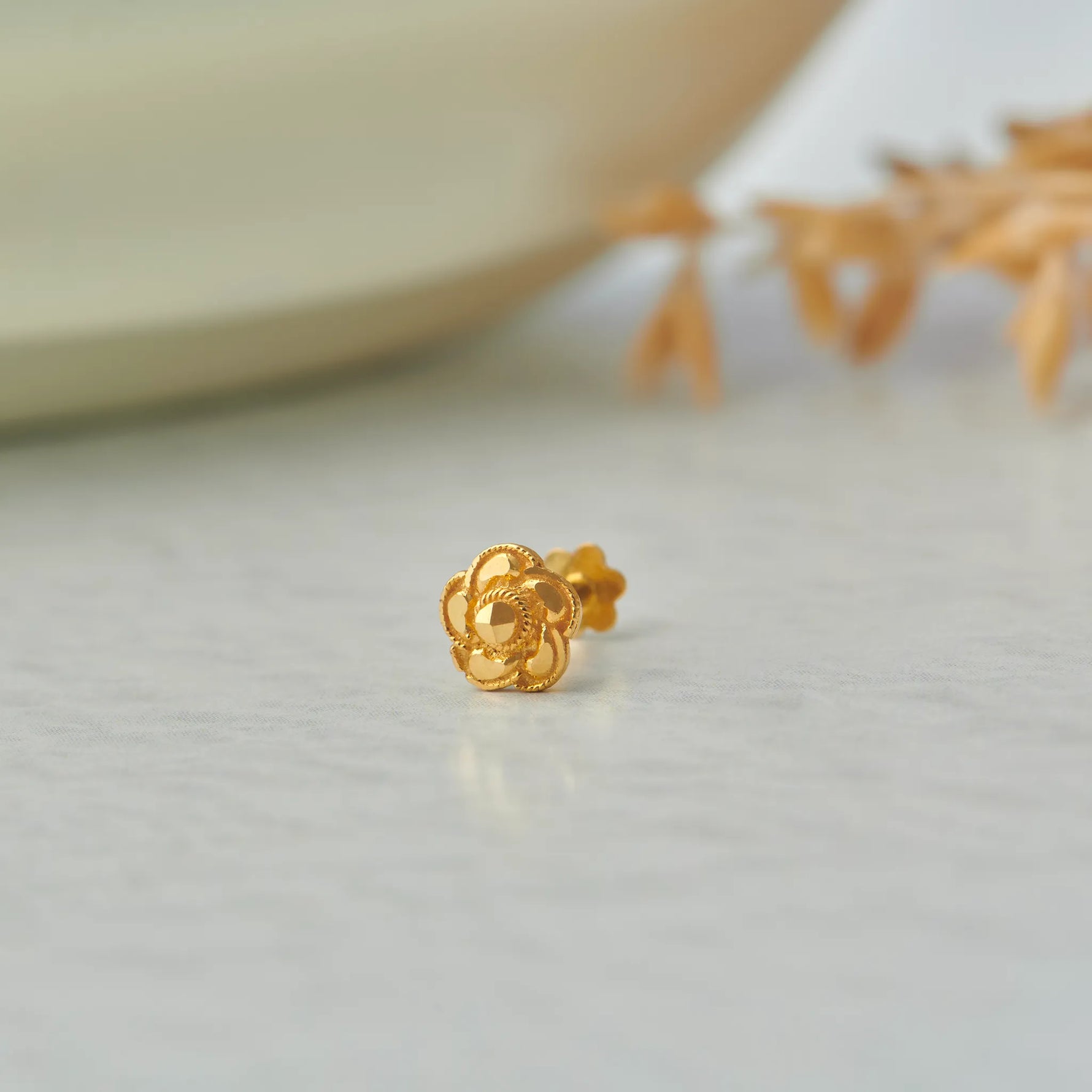 Close up of floral 18K gold nose pin placed on a light textured surface with soft decor elements highlighting the intricate design