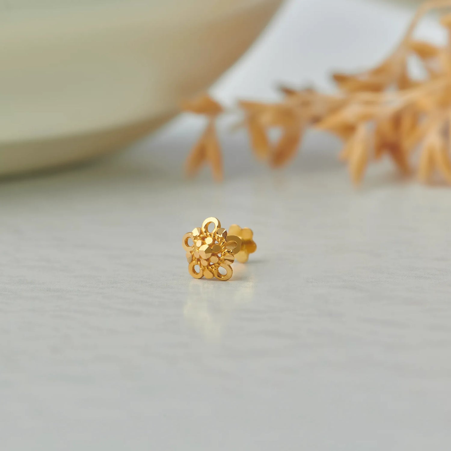 Close up of floral 18K gold nose pin placed on a white surface with soft decor elements highlighting intricate detailing