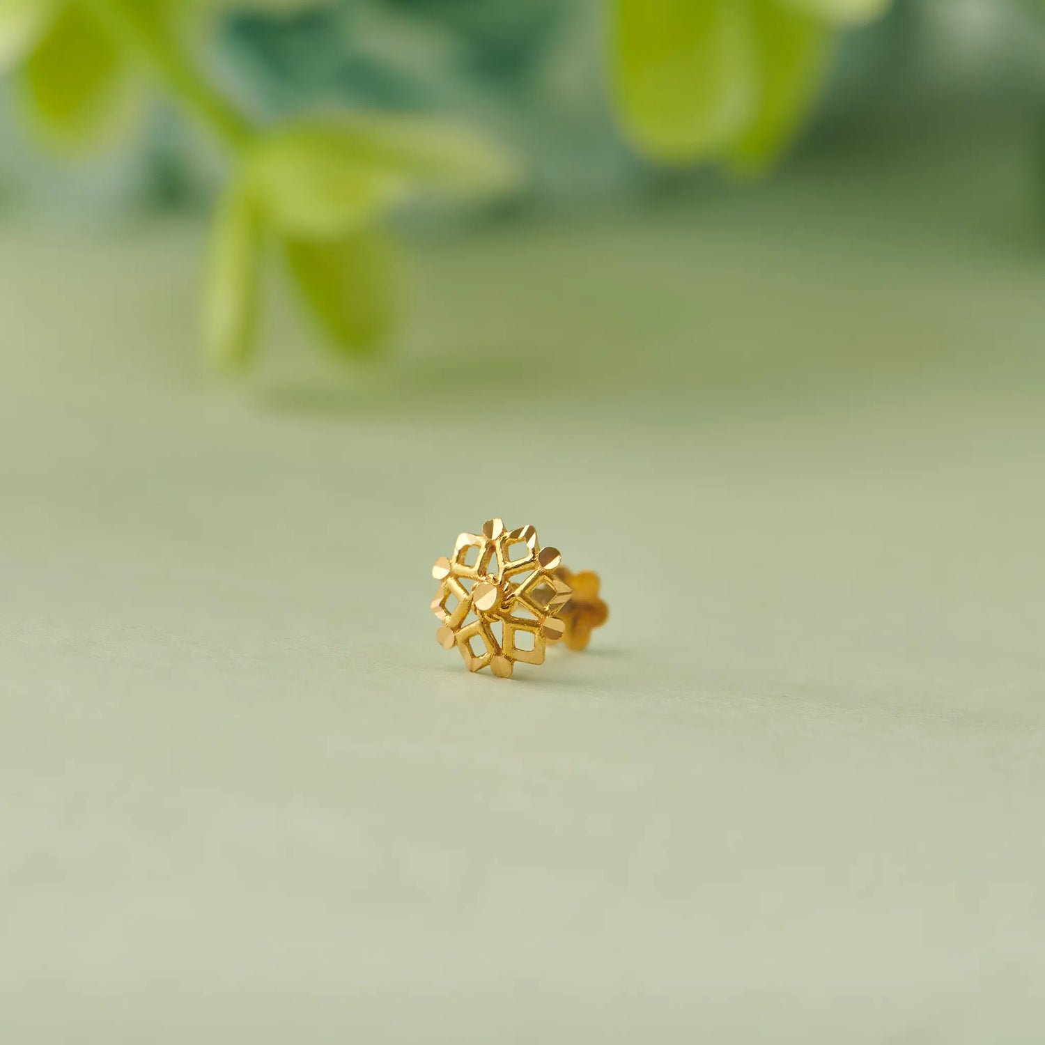 Close up of floral 18K gold nose pin placed on a soft green surface with blurred leaves highlighting its detailed craftsmanship