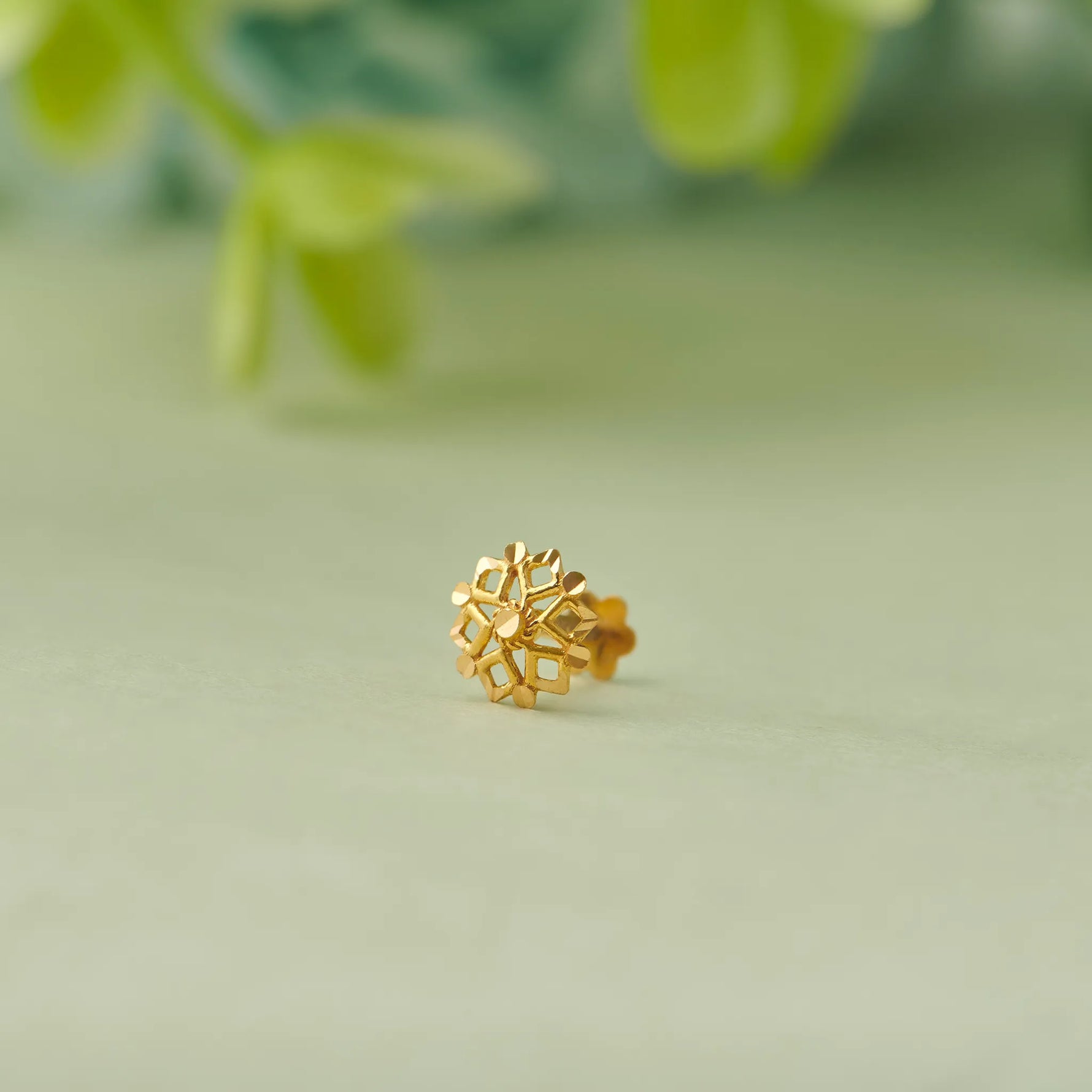 Close up of floral 18K gold nose pin placed on a soft green surface with blurred leaves highlighting its detailed craftsmanship