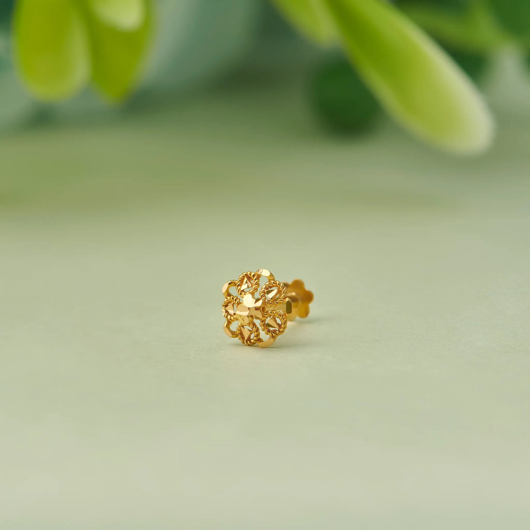 Close up of floral 18K gold nose pin placed on a soft green surface with natural leaves in the background highlighting fine detailing