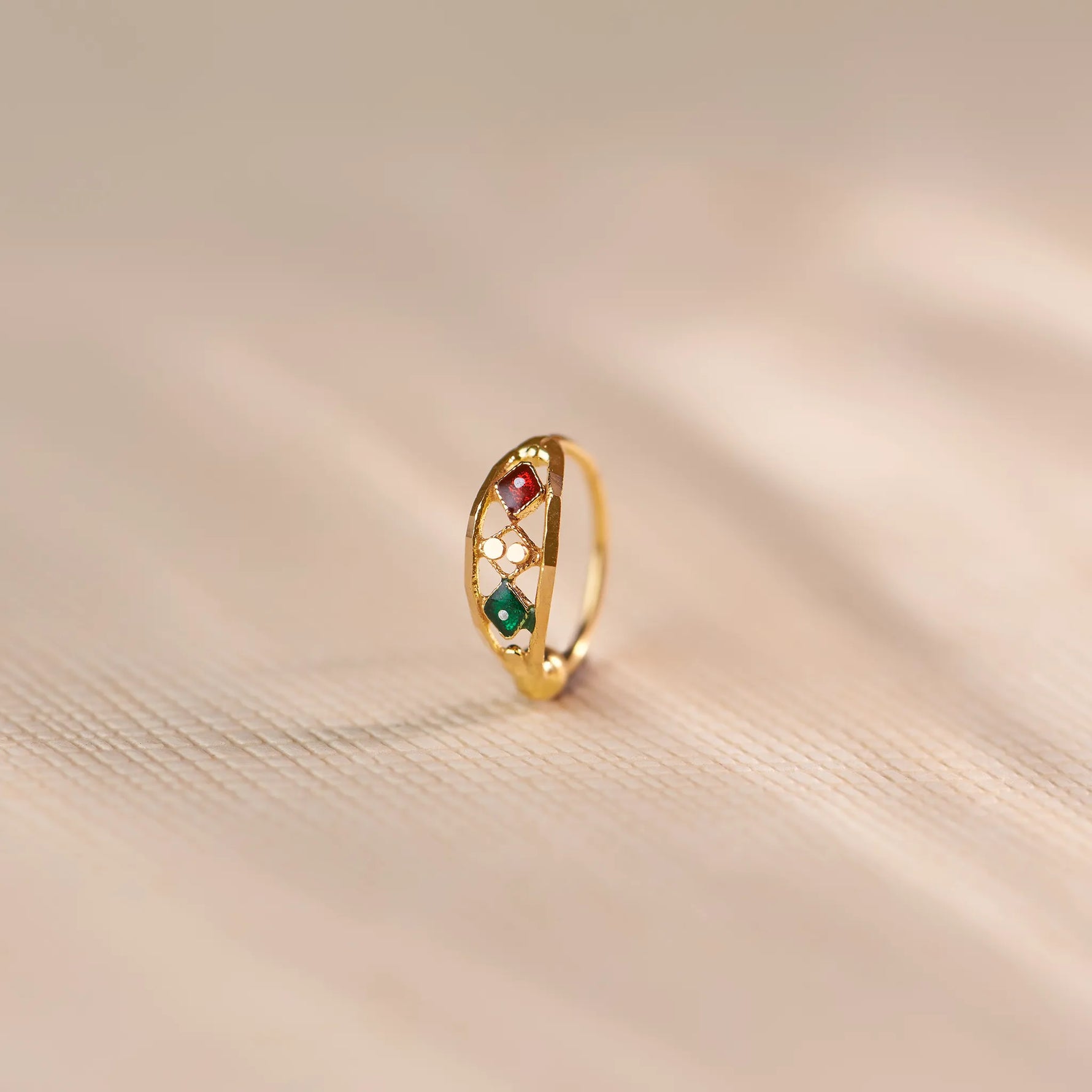 Close up of multicolour stone 18K gold nose ring standing upright on a soft beige fabric background with warm lighting
