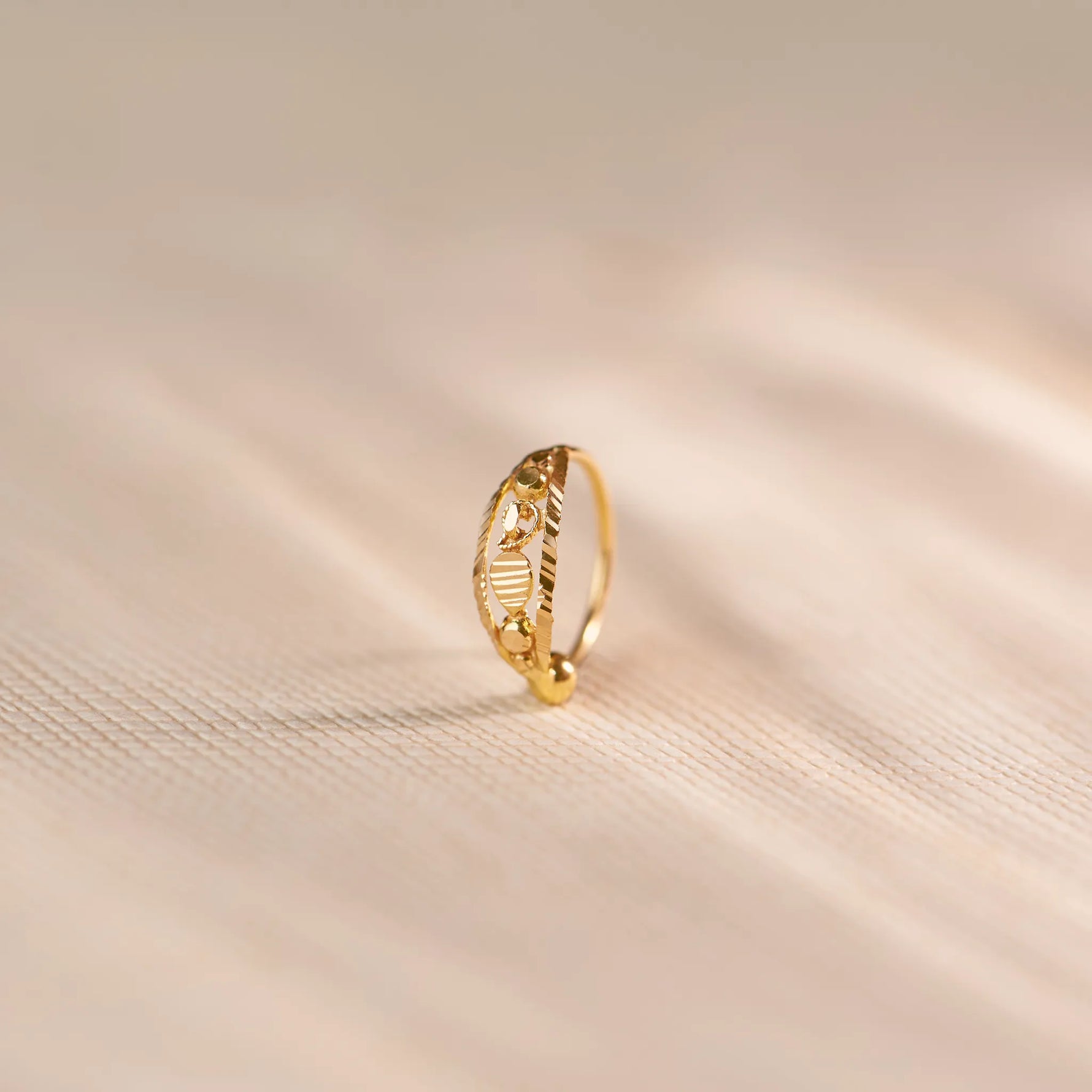 Close up of an 18K gold nose ring with delicate engraved and cutwork details, photographed upright on a soft beige textured fabric with subtle light patterns